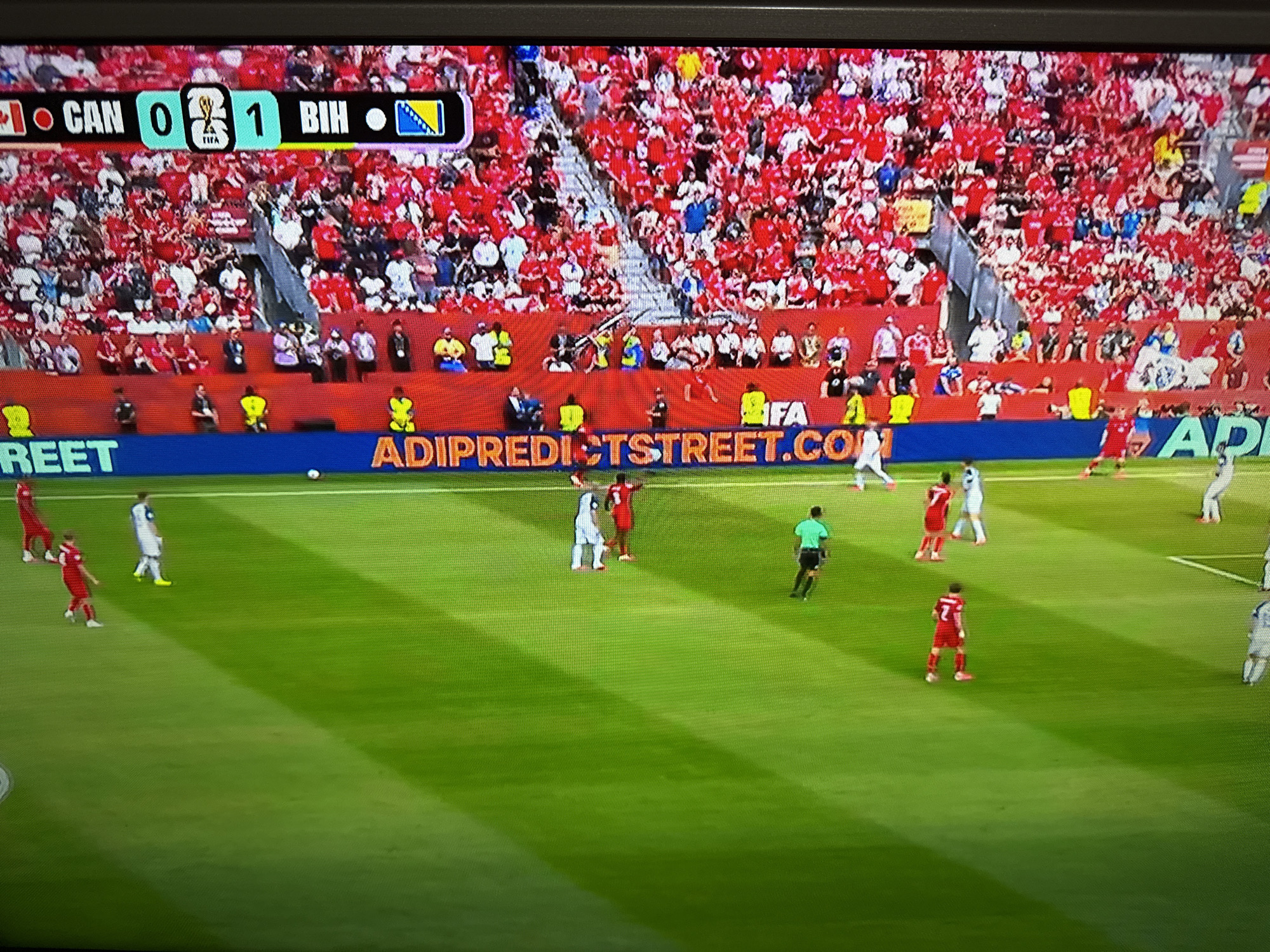 The image shows a soccer match on a bright green field, with a large crowd in the background. The score graphic at the top of the screen indicates CAN 0 - 1 BIH, suggesting a match between Canada and Bosnia and Herzegovina. The stadium is filled with fans, predominantly wearing red. On the field, players in red and white uniforms are visible, some engaged in the game. A blue banner with white text, including "ADIPREDICTSTREET.COM", runs along the lower edge of the field. The lighting suggests it is daytime, possibly late afternoon, given the long shadows cast across the grass. The specific location of this match is not identifiable in the image, nor is there any indication it is in Targovishte, Bulgaria.