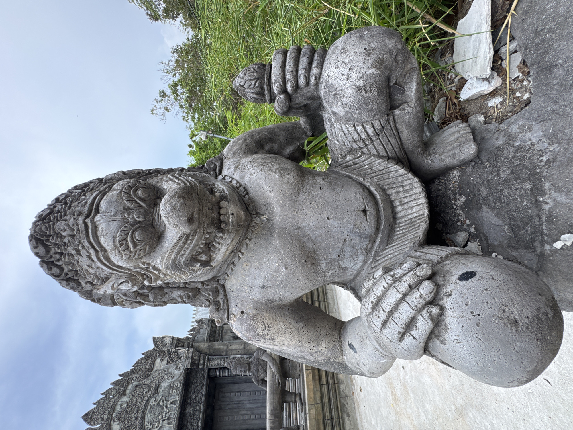 The image shows a stone statue of a mythical creature in a squatting position, holding an object in its right hand. The statue is grey and appears weathered. Behind the statue, there is a structure that resembles a temple with intricate carvings, and lush green vegetation. The sky is overcast. The statue is centrally located in the frame, with a building structure to its left and vegetation to its right. The ground appears to be paved with concrete and scattered with debris. The statue's facial features are prominent, with a fierce expression, sharp teeth, and a broad nose. It is wearing a headdress and a skirt-like garment. The object in its hand appears to be a mace or club.