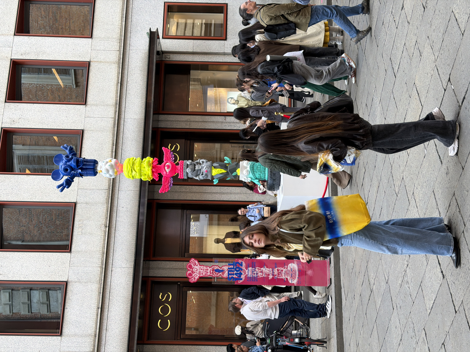 An outdoor urban scene depicts a bustling pedestrian street in Milan, Italy, lined with multi-story buildings and storefronts. The image is rotated 90 degrees counter-clockwise.
In the center of the frame stands a tall, colorful art installation resembling a totem pole, constructed from stacked, stylized figures and abstract shapes. The colors include vibrant blue (at the top, a figure resembling a Mickey Mouse character), white, yellow, neon green, pink, and gray.
Numerous people are visible, walking and standing along the street. In the foreground, a woman with long brown hair wears an olive-green jacket, a light-colored top, and wide-leg blue jeans. She carries a yellow and blue tote bag with the visible text "乾酪" and "ARERA" (or similar). Other individuals are seen further back, some appearing to window shop along the storefronts, while others walk past. One man in the background looks upwards.
The buildings feature multiple windows with dark frames, reflecting the surroundings. A prominent shop entrance displays the brand name "COS" in yellow letters. Next to the main art installation, a tall, rectangular red sign also features a stylized figure and some text including "STILL" and "492", likely pertaining to an art collection or event. The ground is paved with stone, suggesting a pedestrian-friendly area. The scene is brightly lit, indicating daytime. Bicycles or scooters are parked near the bottom left of the frame.