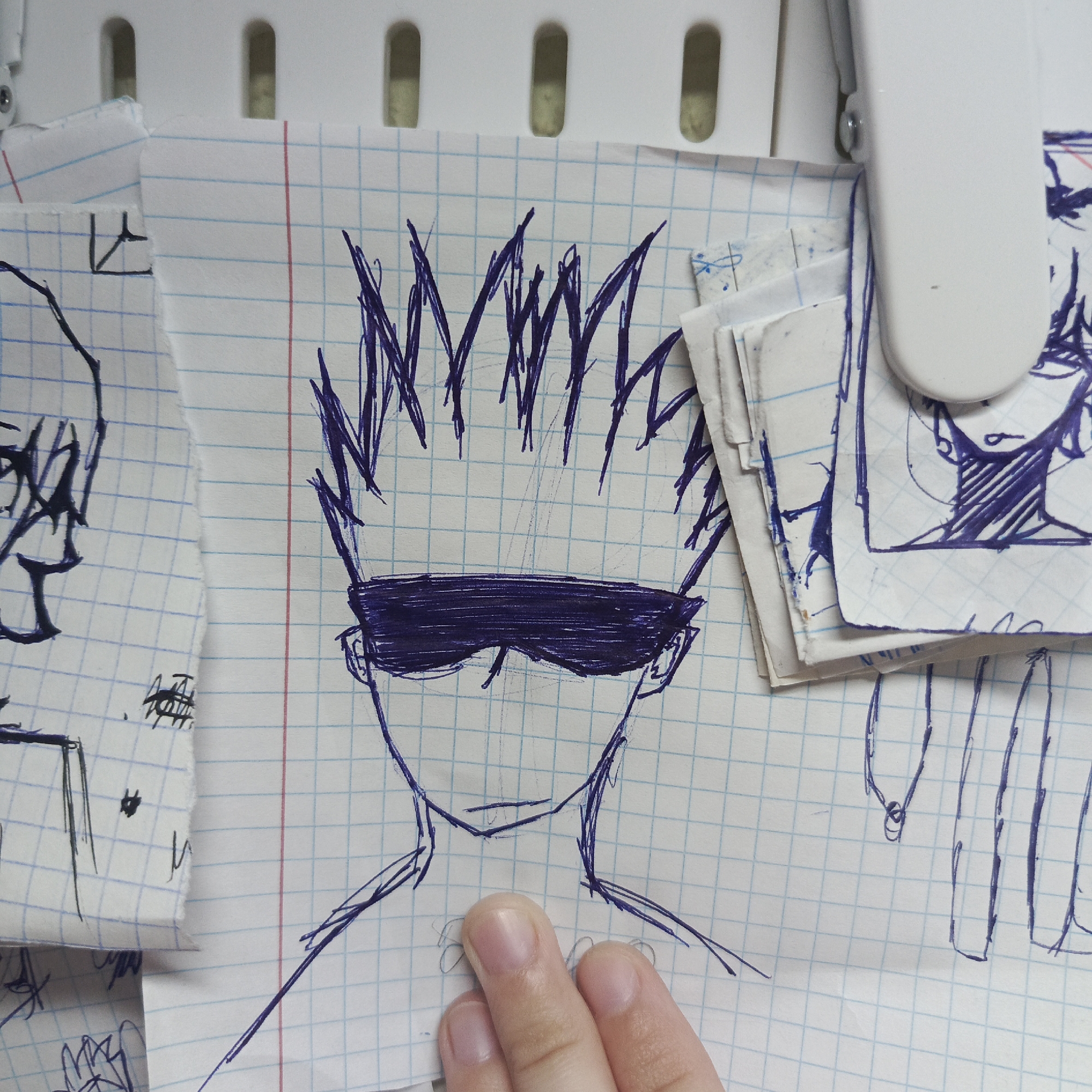 A drawing on graph paper depicts a person with spiky hair and dark glasses. The drawing is done in blue ink and shows only the head and upper torso. The drawing is attached to a white surface, possibly a refrigerator or whiteboard, along with other drawings on graph paper. A human hand is holding the drawing. The location is unknown.