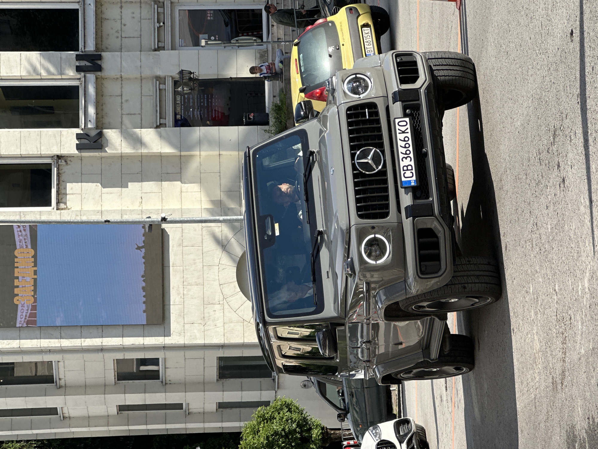 The image shows a gray Mercedes-Benz G-Class SUV parked on a city street with Bulgarian license plate "CB 3666 KO". A small yellow car is parked behind it. People are visible on the steps near the building. The building has the letters "KK" on the front and a sign saying "CINELAND". The location appears to be outside Cineland cinema in Veliko Tarnovo, Bulgaria.