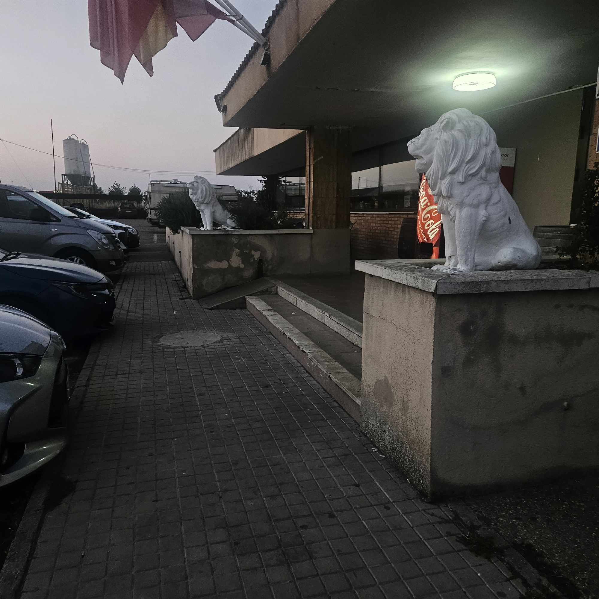 The image depicts an outdoor scene, likely in the early morning or late evening, given the dim lighting and overcast sky. In the foreground, a paved walkway made of dark square tiles leads towards the left side of the frame. Several vehicles, including cars and a van, are parked along the walkway. Two white stone lion statues are prominently featured in the image. One is positioned on a raised concrete platform to the right, and another is visible further down the walkway to the left.
Behind the lion statue on the right, a building with a covered entryway is visible. The building's facade appears to be made of brick and concrete. A large, illuminated Coca-Cola advertisement is mounted on the building's exterior, partially obscured by the lion statue. The entryway is lit by a circular overhead light fixture.
In the background, beyond the parked vehicles, there are some industrial structures, including what appear to be grain silos, and trees. Flags, one red and yellow, are flying from a pole above the building. The overall impression is one of an establishment that may be closed or preparing to open for the day.
The location is identified as Becilla de Valderaduey, Spain.