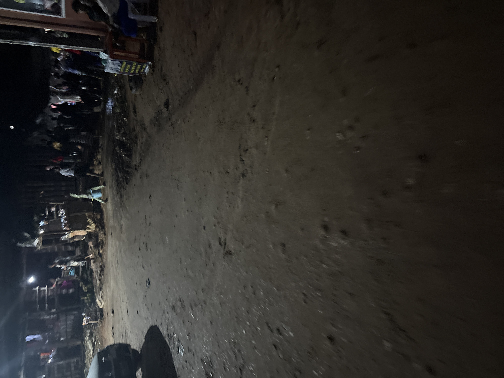The image captures a dimly lit, outdoor scene in Onitsha, Nigeria. The ground is unpaved and appears to be a mixture of dirt and mud, with scattered debris. On the left side of the frame, the rear of a dark-colored vehicle is visible.
In the background, several makeshift stalls or shops are set up, suggesting a market or informal commercial area. Structures appear to be constructed from wood and corrugated metal. A group of people are gathered in the middle ground, their forms partially obscured by the low light. Some individuals are standing, while others appear to be seated or engaged in activities within or around the stalls.
A brightly colored sign, possibly advertising financial services, is propped up on a red stand on the right side of the frame. The sign displays text that is partially legible, mentioning "WITHDRAW DEPOSIT TRANSFER" and "ANYTIME PAY BILE". Two individuals are seated on a white plastic chair in front of this sign, facing away from the camera. The overall atmosphere suggests a bustling, albeit somewhat chaotic, nighttime scene in a developing urban environment.