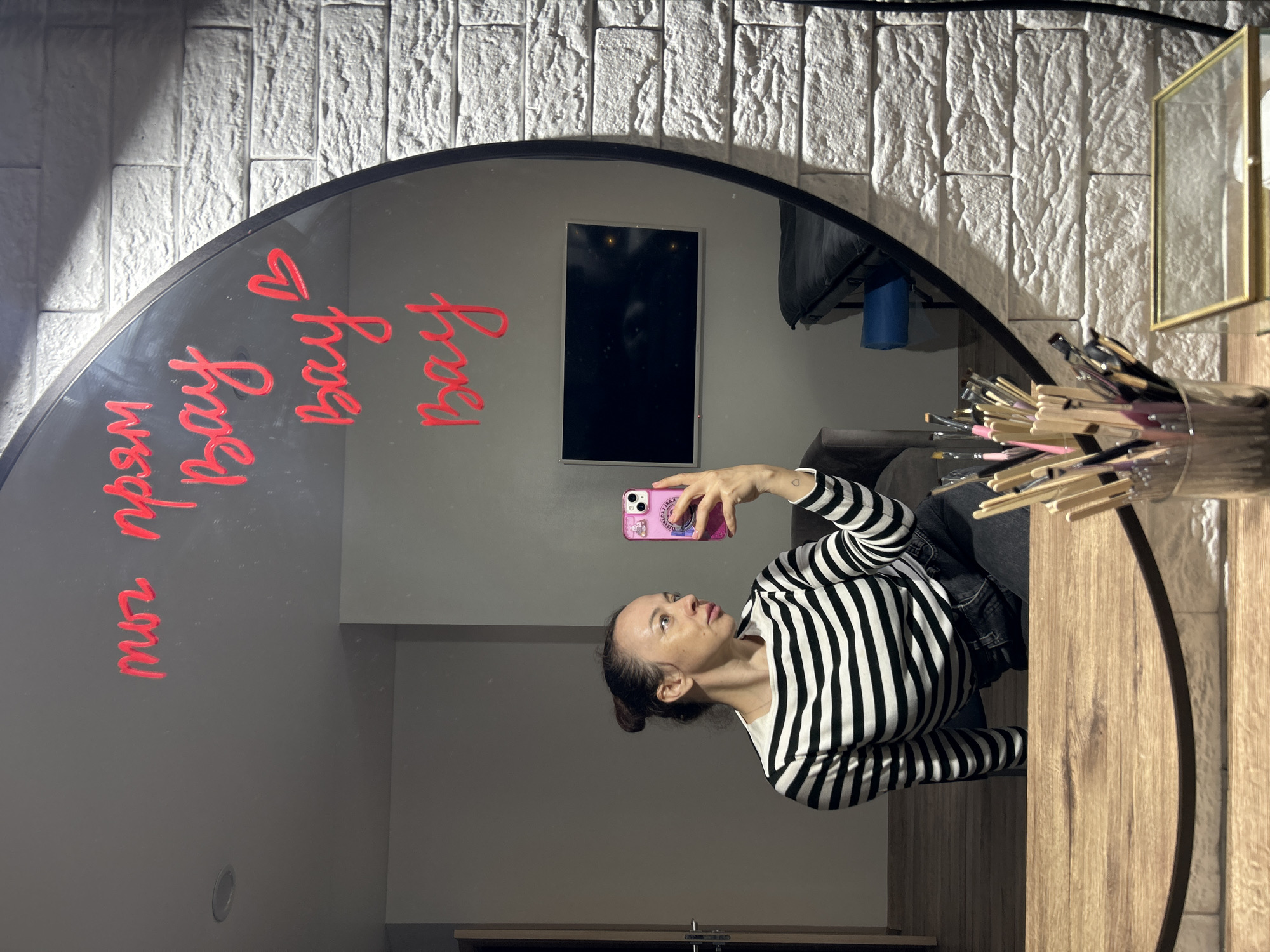 A woman is captured taking a mirror selfie in an indoor setting. She is holding a pink smartphone with a decorated case and is wearing a black and white striped long-sleeved top with dark trousers. Her dark hair is pulled back, and she looks intently at her phone's screen.
The central subject of the image is a large, arched mirror with a black frame. Reflected in the mirror is the woman herself, along with various elements of the room. A distinctive feature on the mirror's surface is red script writing, which clearly spells out "Bay Bay Bay ❤️" (with a heart symbol) and "wow wow wow" (reversed and partially obscured).
The room's decor, visible in the mirror's reflection, includes a textured white brick-patterned wall. Also reflected are a black-screen flat-panel television, a dark sofa or chair, and a wooden surface that appears to be a desk or vanity. On this wooden surface, a clear cylindrical container holds a large collection of makeup brushes, and a gold-framed object is visible next to it. In the background, a blue cylindrical object and part of what appears to be lighting equipment are also reflected.
The activity taking place is a mirror selfie, suggesting the woman is either preparing for an event or simply capturing a moment in a space that resembles a personal vanity area or a beauty studio. The overall atmosphere appears neutral. The location context provided is Chaikovsky, Russia.