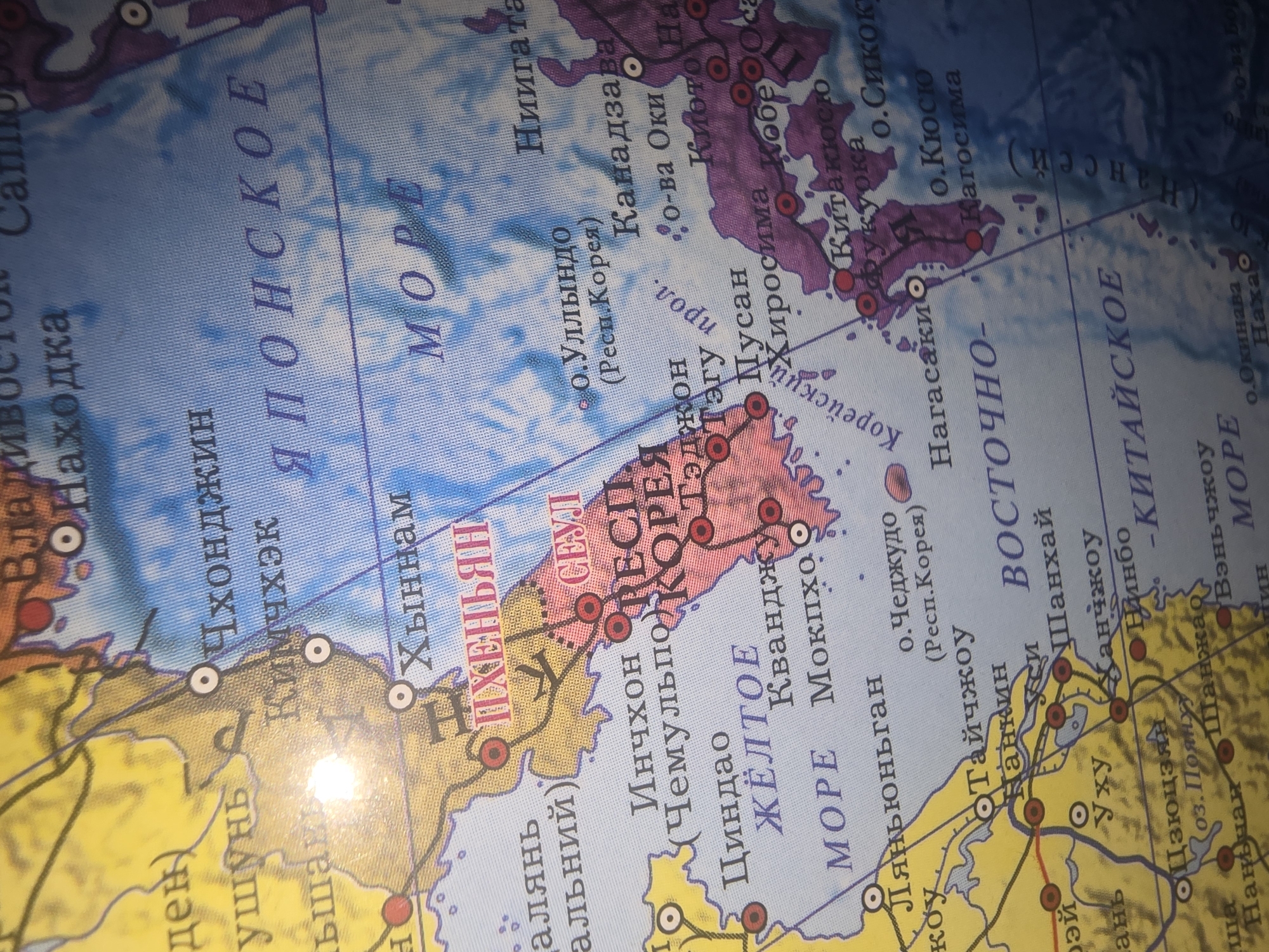 The image is a close-up photograph of a globe, focusing on East Asia and the surrounding seas. The globe displays various geographical locations, including countries, cities, and bodies of water, marked with text in Russian. Prominently visible are the Korean Peninsula (divided into North and South Korea), Japan, parts of China, and the Sea of Japan (labeled as Японское море) and the Yellow Sea (labeled as Желтое море). Several cities are labeled in Cyrillic, such as Pyongyang (Пхеньян), Seoul (Сеул), Incheon (Инчхон), and various Japanese cities like Nagasaki (Нагасаки) and Hiroshima (Хиросима). The colors on the globe represent landmasses in yellow and light brown, and water bodies in shades of blue. The lighting in the image suggests an indoor setting, with a bright glare reflecting off the globe's surface, indicating the presence of a light source. There are no people, animals, or specific actions depicted; it is purely a static representation of a map on a globe. The city of Ulan-Ude, Russia, is not directly visible or labeled in this particular section of the globe.