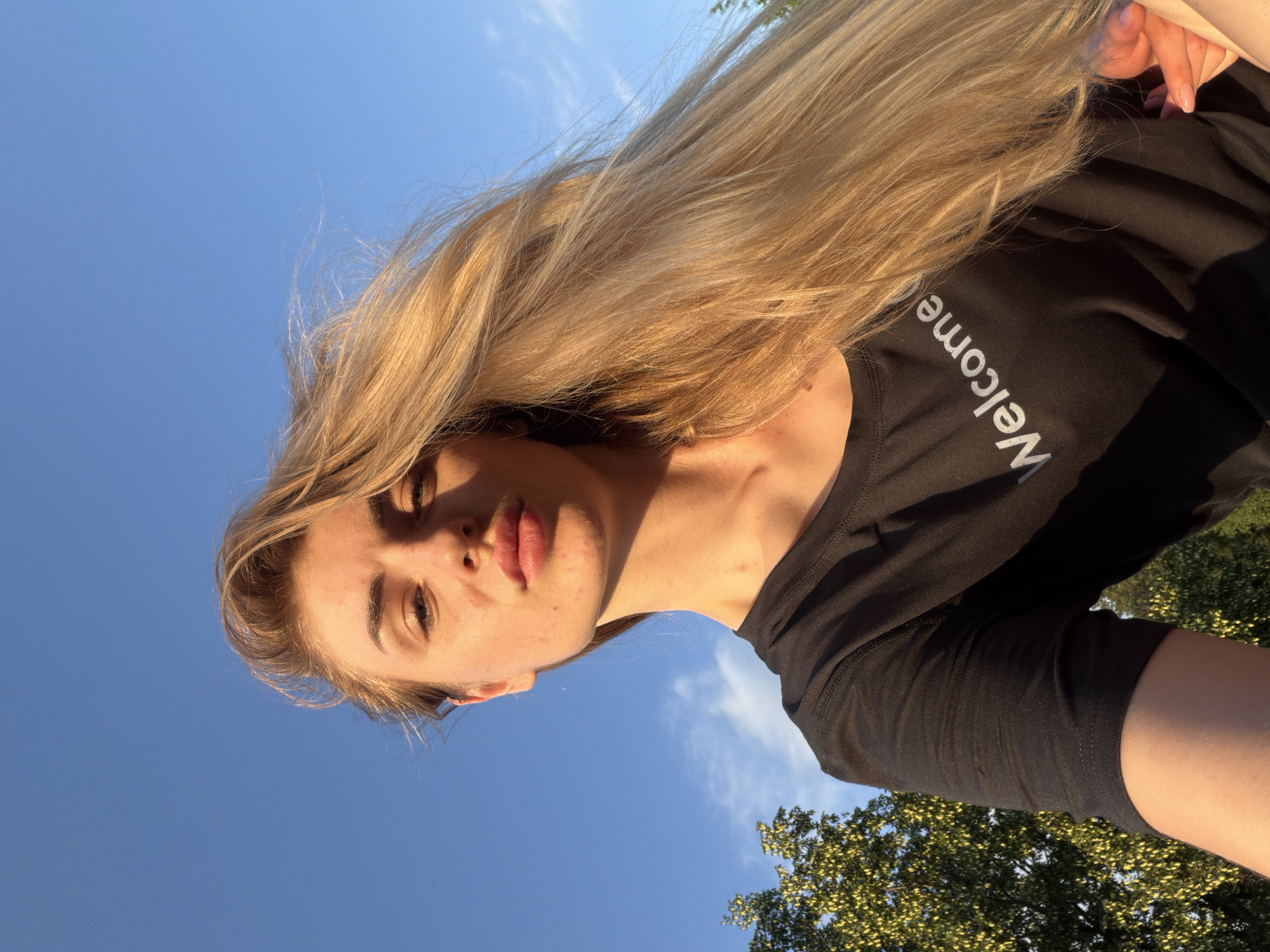 A young woman with long blonde hair is captured in a low-angle selfie against a clear blue sky. She is wearing a black t-shirt with the word "Welcome" printed in white. The setting appears to be outdoors, with trees visible on the left side of the frame. The golden hour lighting casts a warm glow on her face and hair.