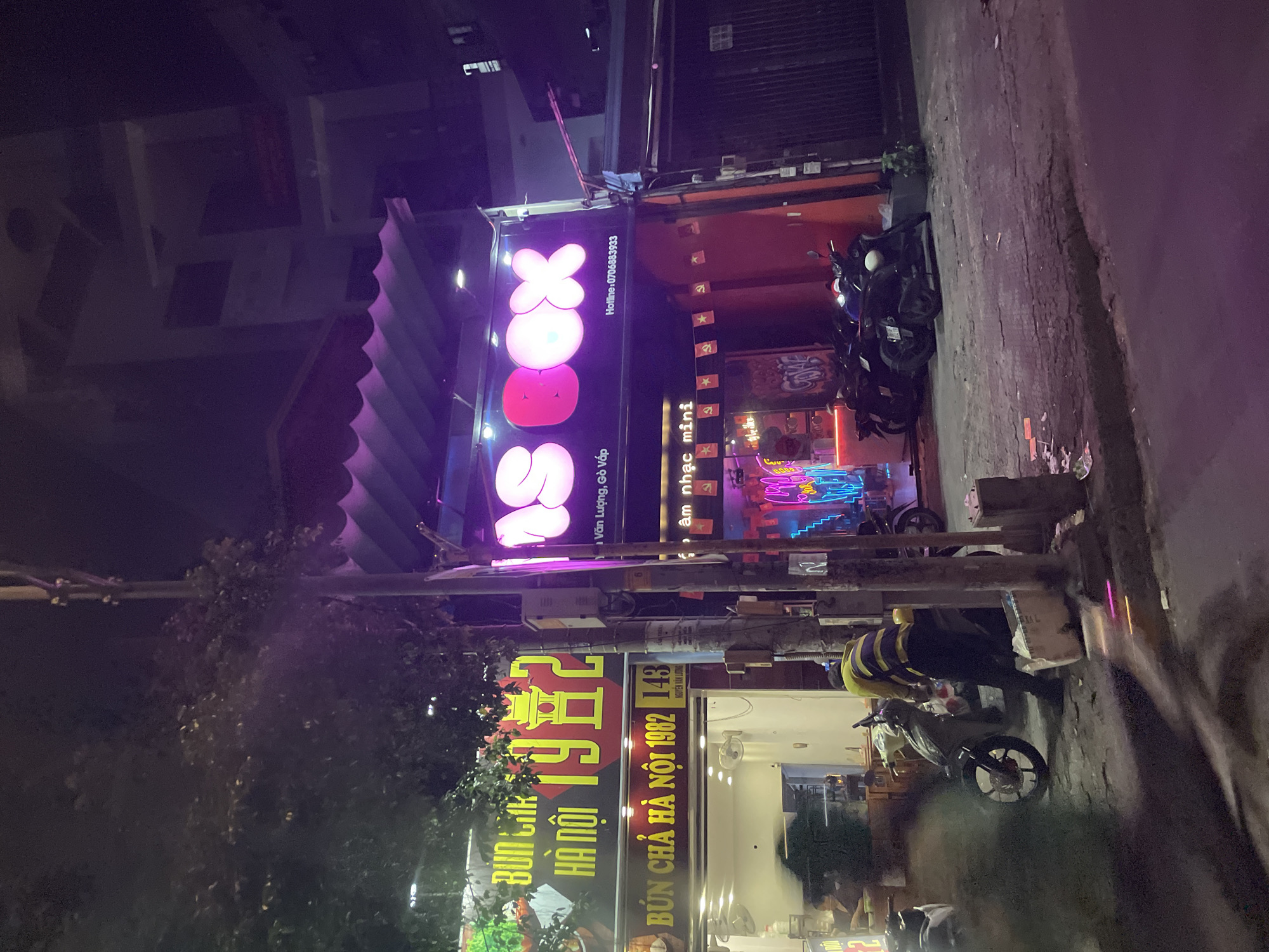 The image shows a street scene at night in Thuận An, Vietnam. On the left, a brightly lit sign displays "BUN CHIK HA NOI 1982" and below it "BUN CHA HA NOI 1982 143 NGUYEN VAN LUONG". A person in a yellow and purple striped shirt is standing next to a parked motorcycle. In the background, a neon sign reads "SBOX" in pink lettering, with a phone number "0706883933" beneath it. Further down the street, another neon sign with a pink and blue glow displays text that is partially obscured. Several motorcycles are parked along the sidewalk. The environment appears to be a commercial street with shops and businesses.