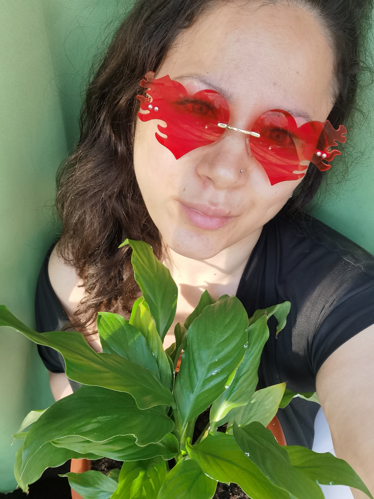 A medium-angle, close-up shot depicts a woman with dark, curly hair wearing red heart-shaped sunglasses adorned with flame-like extensions. She is holding a potted plant with vibrant green leaves in front of her, possibly a Peace Lily. She is wearing a dark-colored, off-the-shoulder shirt. The background is a solid, light green color. The location, Chaikovsky, Russia, is not evident in the image.