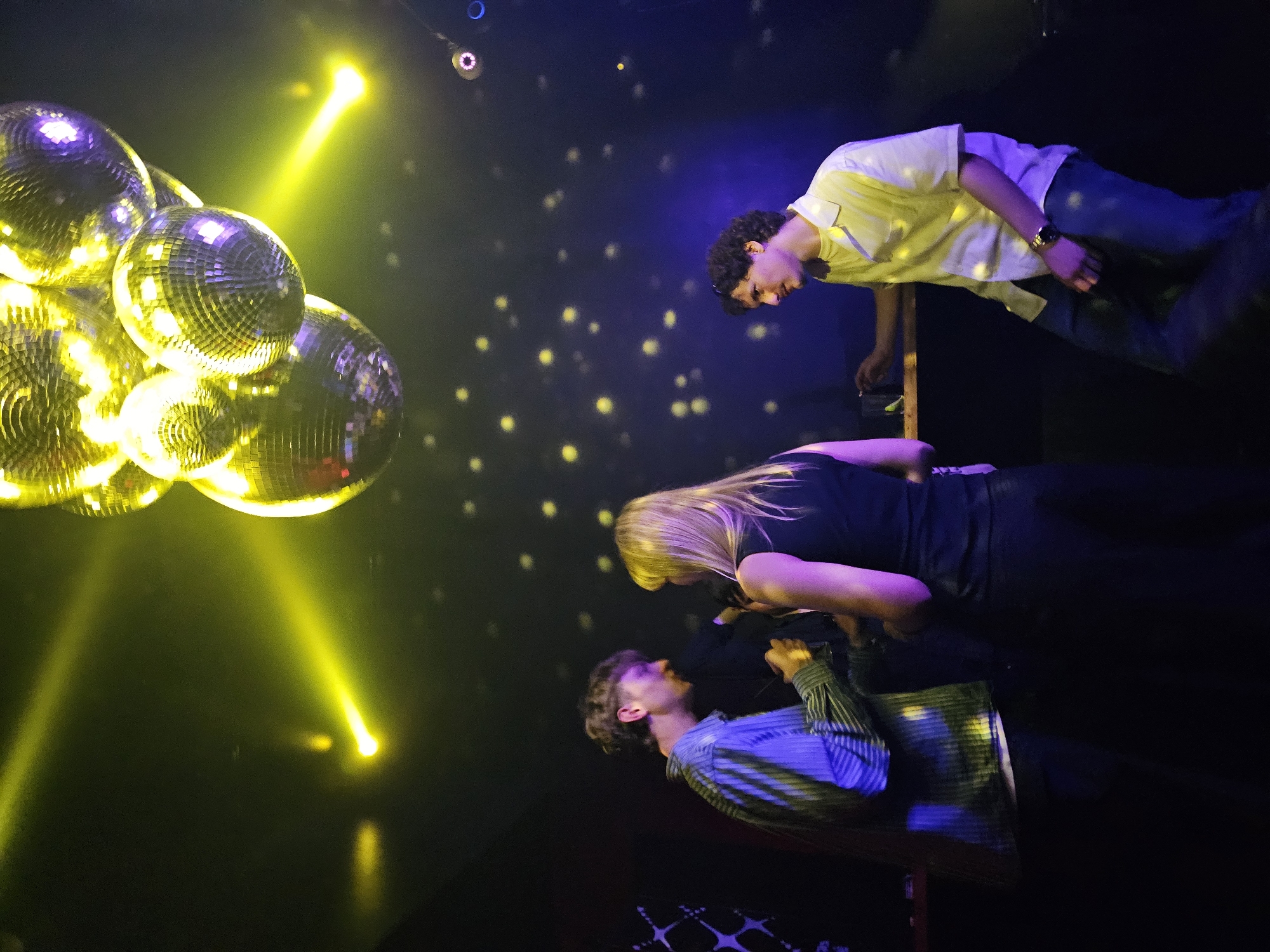 Three individuals stand on a raised platform under a disco ball, illuminated by yellow spotlights and scattered star-like lights, possibly within a nightclub setting. The person on the left wears a blue striped shirt, while the person in the middle has blonde hair and wears a black shirt. The person on the right wears a yellow shirt and blue jeans and leans on a stand. The setting is a nightclub or stage. There are no indications the scene is in Paris, France.