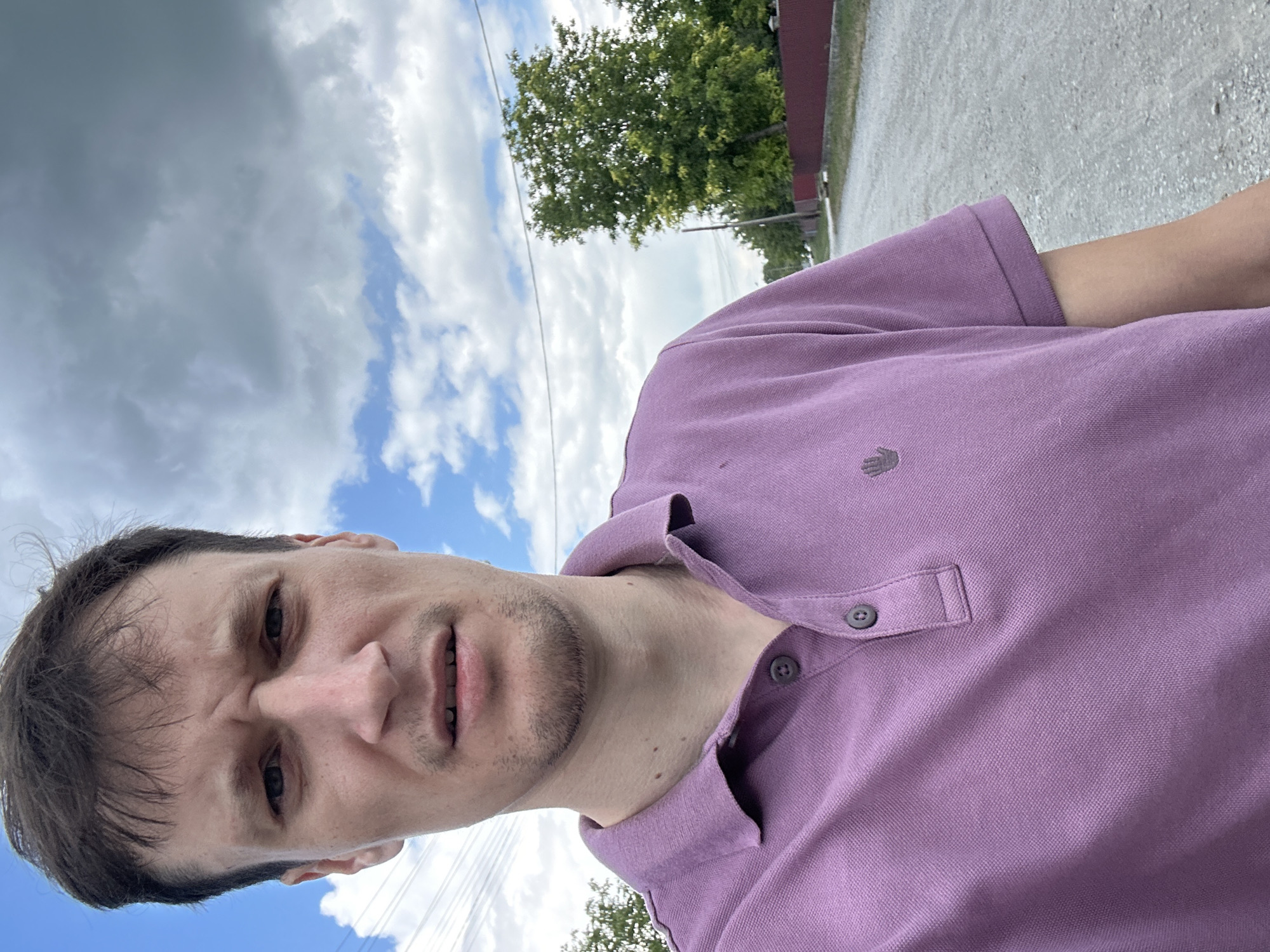 A selfie of a Caucasian male is taken outdoors in what appears to be a rural setting in Makiv, Ukraine. The man has fair skin, light brown hair, and a serious expression. He is wearing a light purple polo shirt with a small grey embroidered hand logo on the left side of the chest. The background shows a grey gravel road, a red fence, green trees, and a cloudy blue sky with power lines visible. The man is looking directly at the camera with his mouth slightly open.