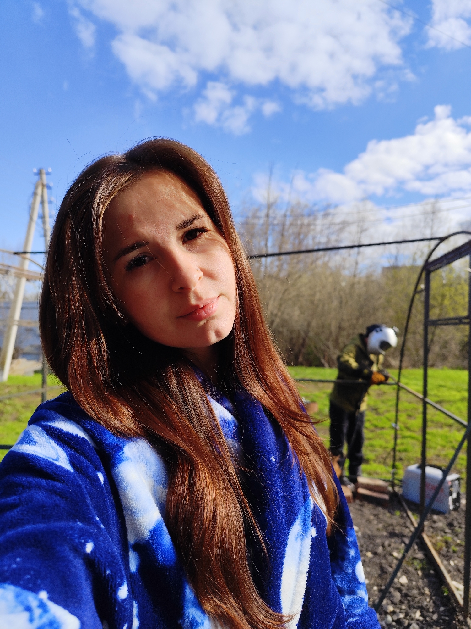 A fair-skinned woman with long brown hair is posing for a selfie. She is wearing a blue and white tie-dye patterned fleece garment. Behind her is a man wearing a white helmet and a green jacket welding a metal structure. A white container is next to the structure. The background features a view of trees and a bright blue sky with scattered clouds. Power lines and poles are visible on the left side of the frame. The location is Фоки, Russia.