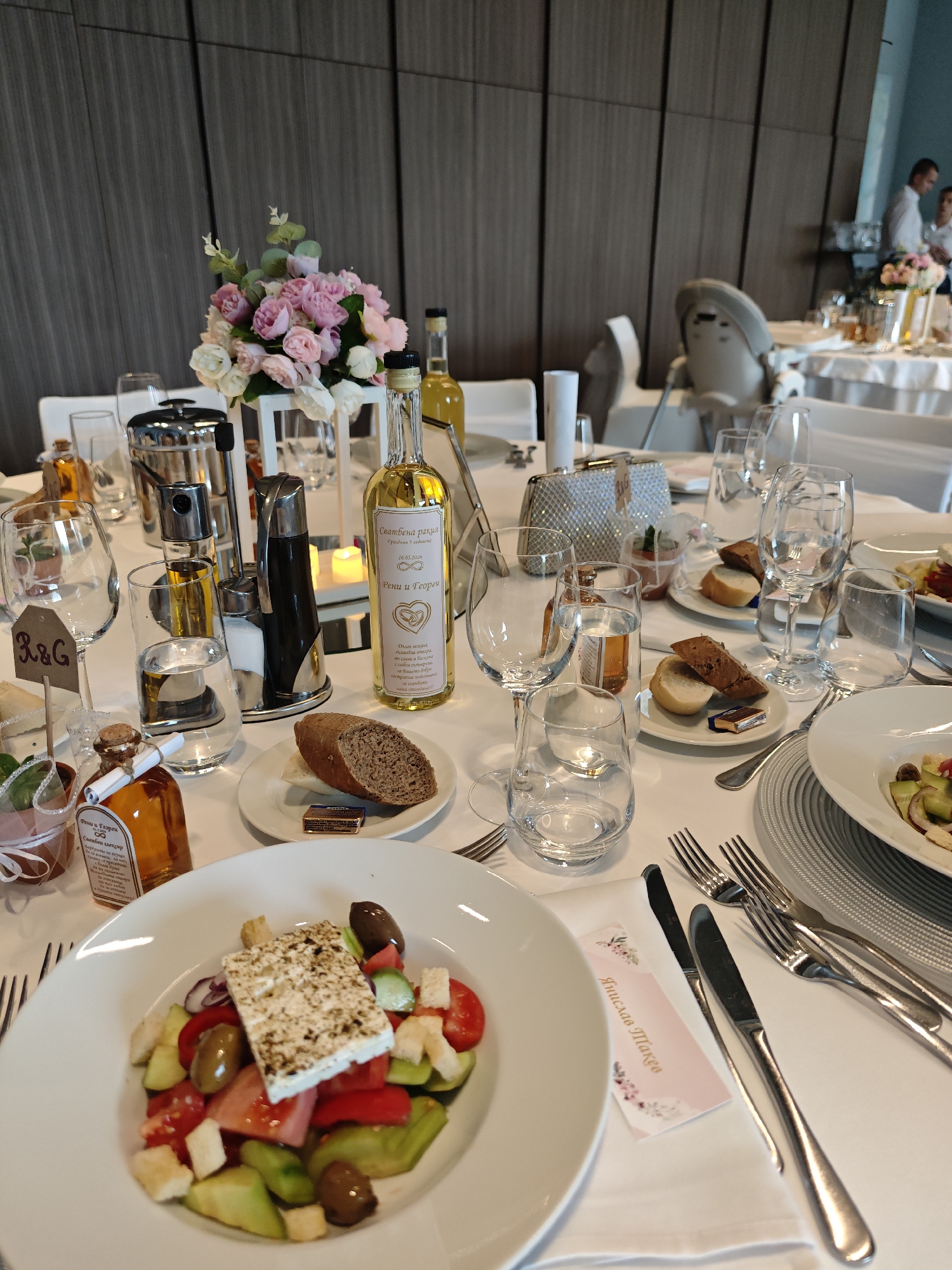 The image captures a wedding table setting. In the foreground, a plate of Greek salad is visible, along with bread, butter, oil and vinegar cruets. The tables are covered with white tablecloths, and the setting includes multiple glasses, forks, and knives. A small card with Cyrillic text, "Янислав Такев" (Yanislav Takev), sits beside the place setting. The table is decorated with a bouquet of pink and white roses in a white lantern-style holder. Bottles of Bulgarian "Rakia" are also included, and a small wedding gift with the initials "R&G" can be seen. In the background, additional tables with white tablecloths and similar settings are visible, along with a high chair and people in the distance. Based on the writing and style of the wedding arrangements, the image is likely taken in Bulgaria, possibly in the city of Sofia.