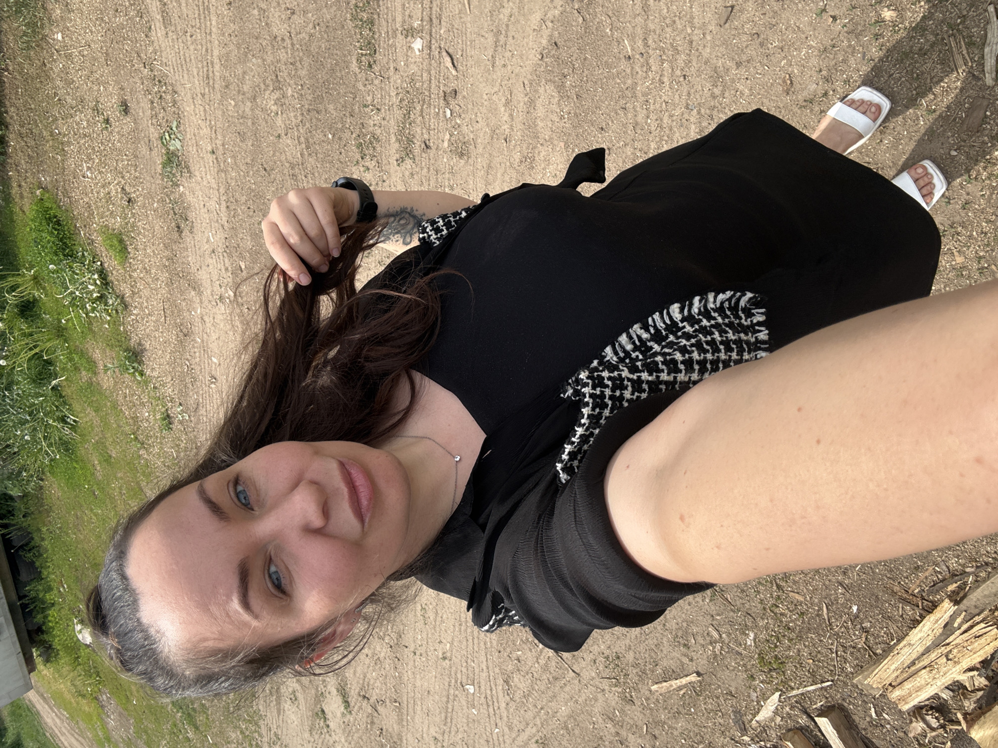 A woman with light blue eyes and brown hair is taking a selfie outdoors. She is wearing a black dress with a black and white patterned jacket and white sandals. She is standing on a dirt path with some grass and scattered pieces of wood. She is wearing a watch on her left wrist, which also bears a tattoo. The photograph appears to be taken in an outdoor location, possibly in or near Chaikovsky, Russia.