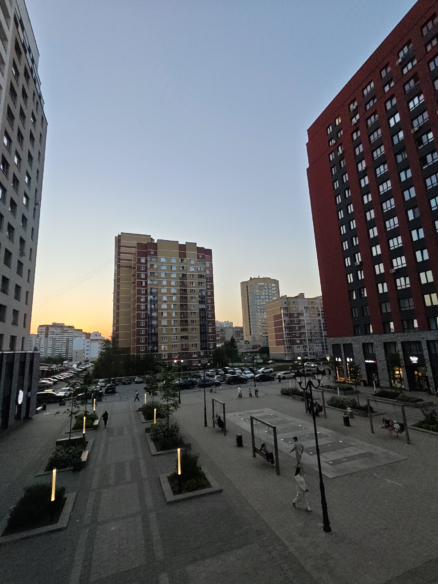 The image depicts an urban landscape in what appears to be Yekaterinburg, Russia. The view is elevated, looking down onto a pedestrian plaza bordered by modern apartment buildings. The buildings are predominantly high-rise, with one red-brick building standing out on the right. A yellow and brown building is located to the center. A paved plaza features several benches, decorative planters, and people. Some are walking, and others are seated or swinging on playground-style swings. Beyond the plaza, a parking lot is filled with cars. The sky is colored in twilight hues, indicating the time of day. Street lamps light the plaza.
