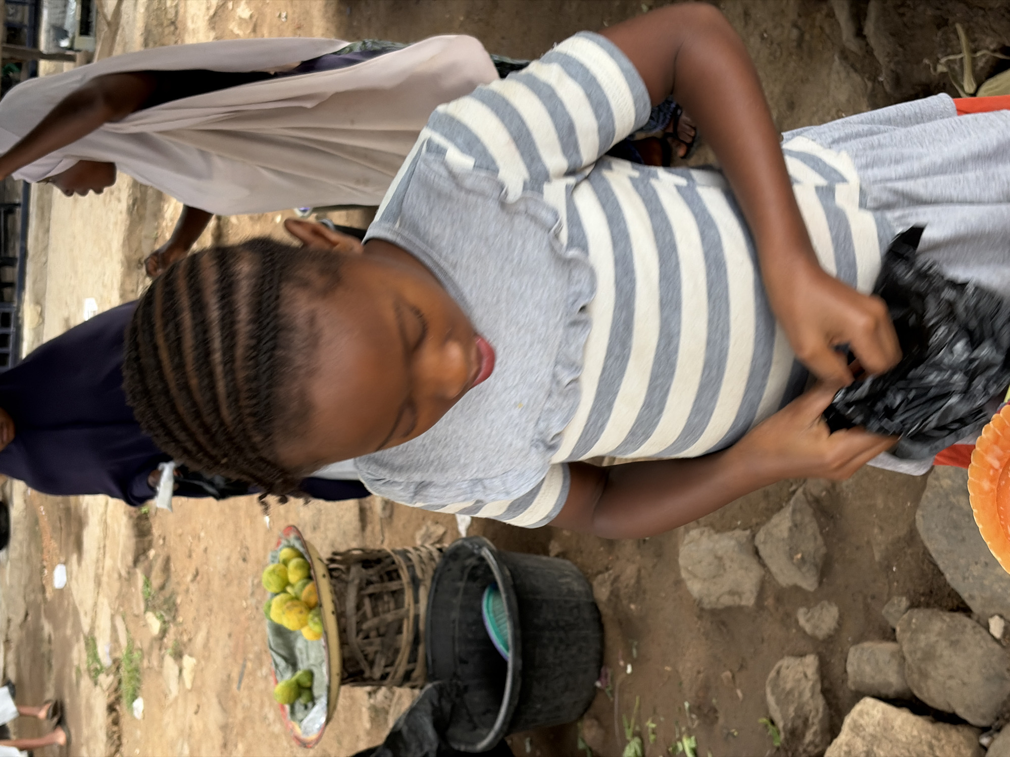 The image captures a bustling outdoor market scene, likely in the city of Jos, Nigeria. In the foreground, a young girl with braided hair is focused on holding a black plastic bag, her hands manipulating its contents. She is wearing a grey and white striped t-shirt with a ruffled neckline, and a grey skirt. Behind her, to the left, a tray of green and yellow fruits rests on a woven basket. To the right, two women are partially visible; one is wearing a light-colored hijab and appears to be engaged in conversation, while the other, wearing a dark head covering, stands behind a stall. The ground is unpaved, with patches of dirt and scattered rocks, indicating an informal market setting. Several black plastic containers are also present in the scene. The overall impression is one of daily commerce and activity within a Nigerian urban environment.