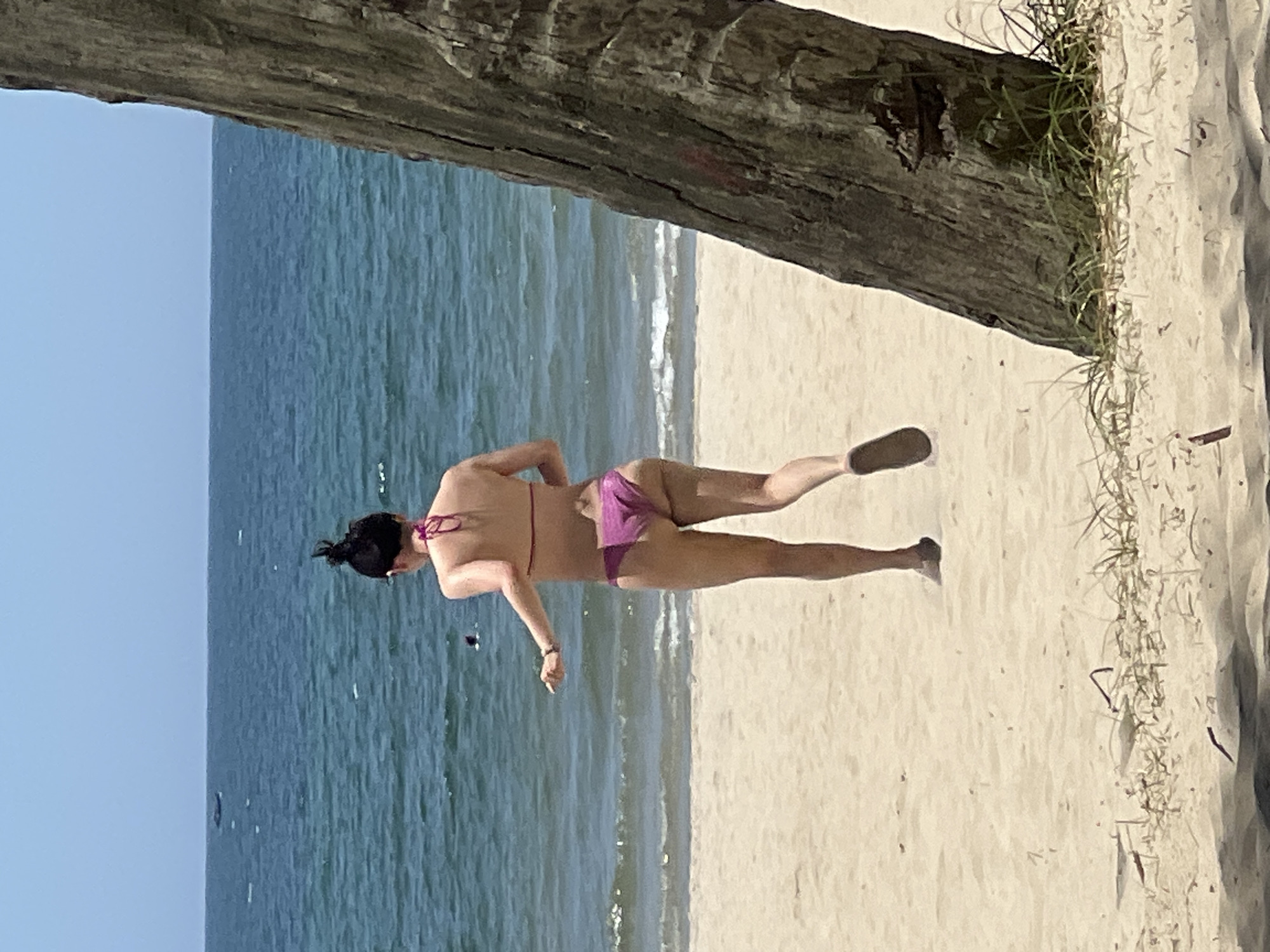 A person wearing a pink bikini is running on a sandy beach with the ocean in the background. The person's back is to the camera, and they have their hair pulled up in a bun. There is a palm tree trunk on the right side of the image. The ocean is blue with some whitecaps, and the sky is clear and blue. The beach is light brown sand with some scattered vegetation. The scene appears to be in Da Nang, Vietnam, a coastal city known for its beaches.