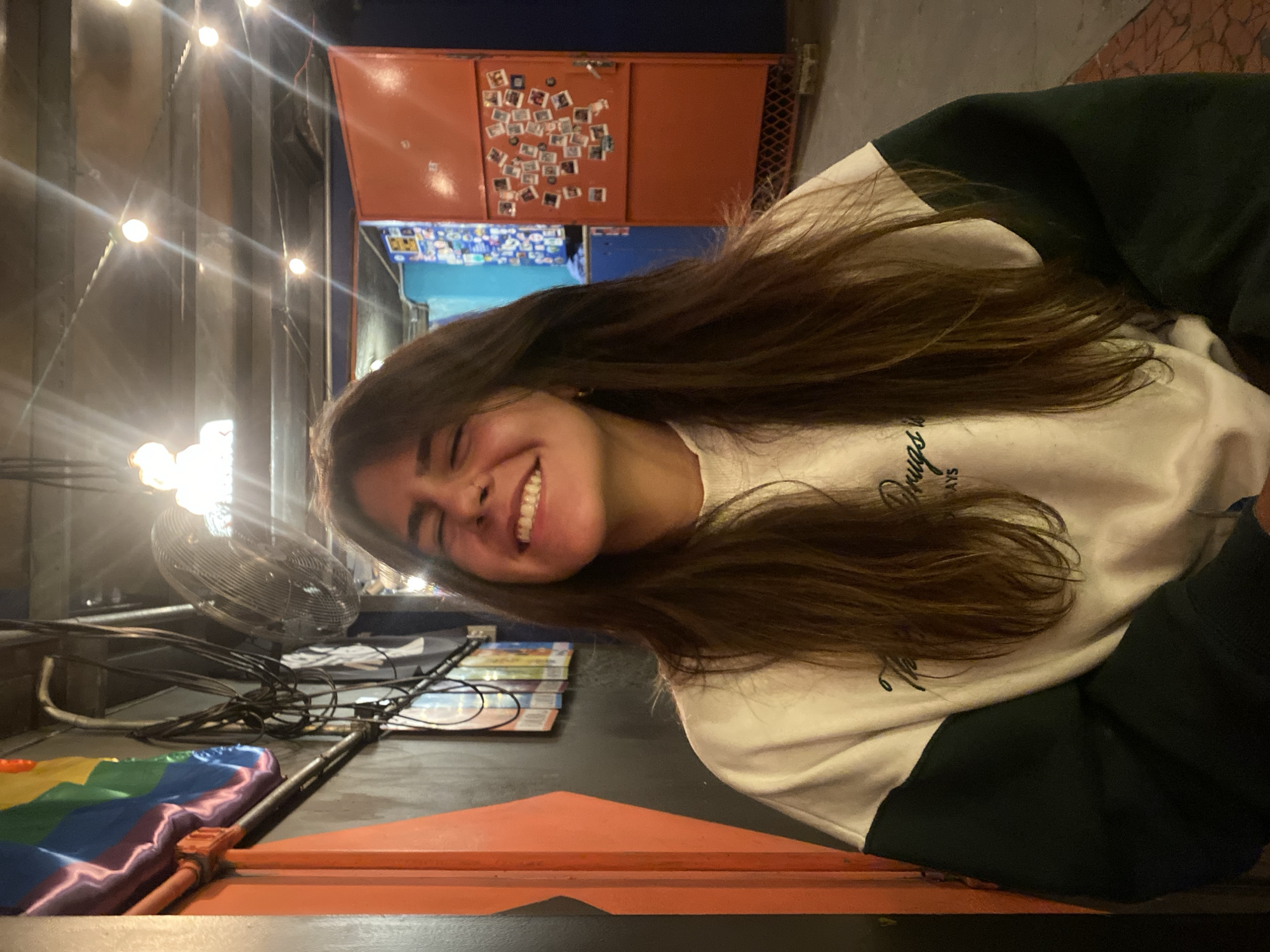 A young woman with long brown hair is smiling in an indoor setting, seemingly at a restaurant or bar. She is wearing a white and dark green sweatshirt. Behind her, there are strings of lights and a metal structure with an orange painted exterior decorated with numerous small square photographs. Further back, there is a blue wall also decorated with photos. To her left, there is a metal fan and a rainbow-colored pad on a surface. The location is likely in the city of São Paulo, Brazil, based on the general ambiance and architectural style.
