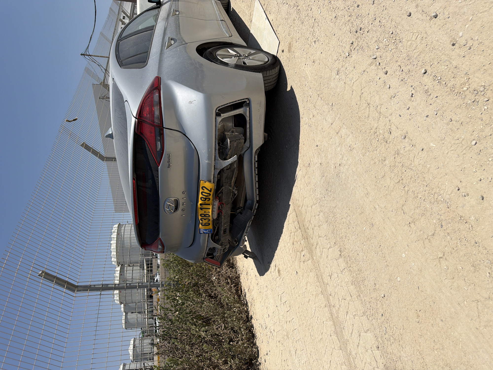 The image shows the rear of a silver Hyundai Ioniq hybrid car with significant damage to its rear bumper and lower body. The license plate, 638-11-902, is visible. The car is parked on a dirt and gravel surface. In the background, there is a chain-link fence, several large cylindrical storage tanks with white conical tops, and some green bushes. The sky is clear and blue. The location is identified as Rishon LeZion, Israel.
