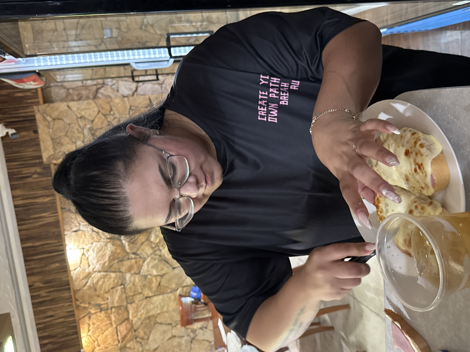 A woman with dark hair pulled back in a bun and wearing glasses and a black t-shirt with pink text is slicing food on a white plate. The plate contains several pieces of bread topped with melted cheese and what appears to be ham. A clear plastic cup filled with a light-colored beverage and ice is to the left of the plate, and another slice of bread with ham is visible to the far left. The woman is wearing a bracelet on her right wrist and a ring on her left ring finger. The background features a stone wall and a wooden wall. The image appears to have been taken indoors, possibly in a restaurant or dining area in Sredets, Bulgaria.
