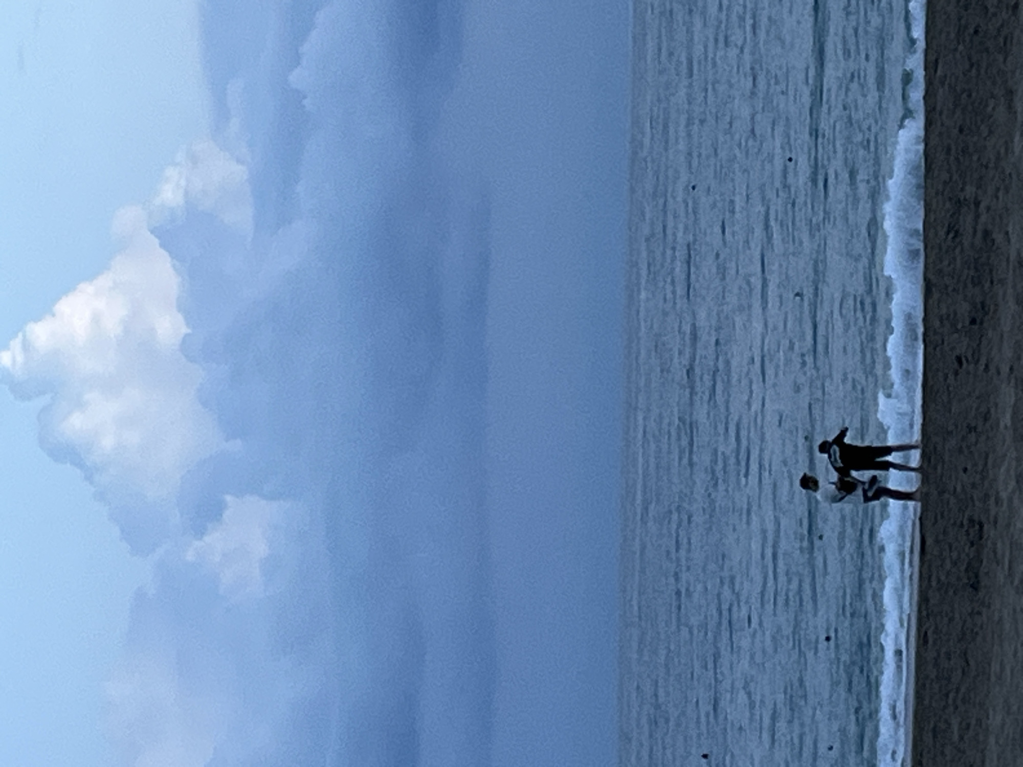Two people stand on a beach at the edge of the water. A large, white cloud formation dominates the left side of the frame. The ocean occupies the center. The beach is on the right. The scene appears to be Alanya, Turkey.