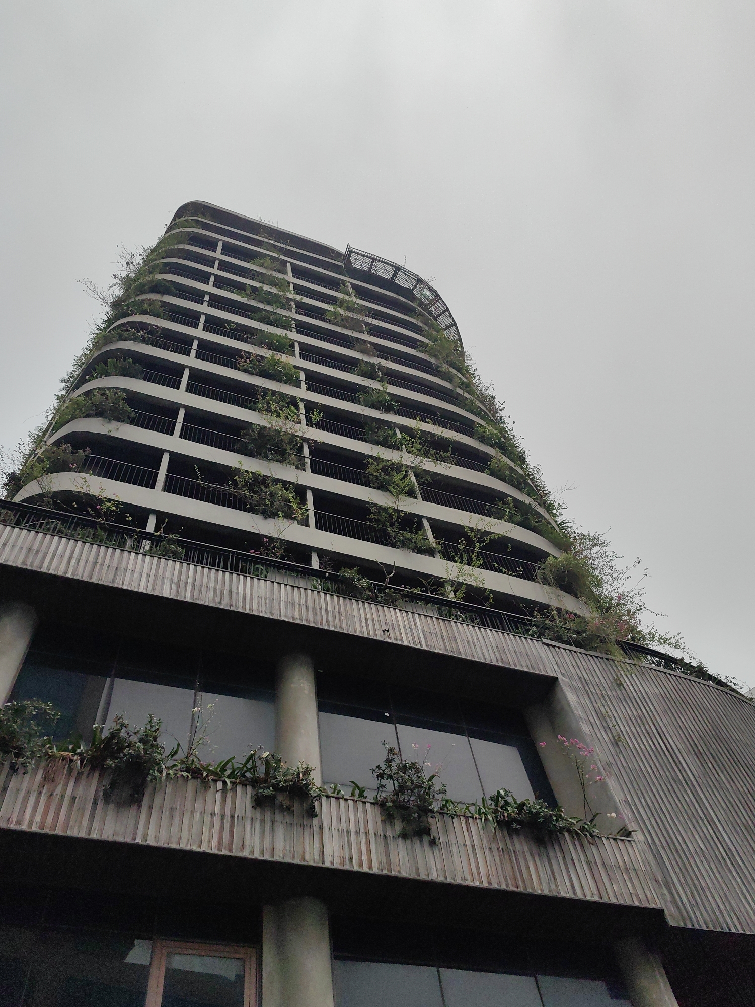 The image depicts a tall building in Bai Chay Ward, Vietnam, characterized by its rounded corners and multiple balconies. Each balcony is lined with greenery, including plants and flowers. The building's lower levels feature large windows and a façade with vertical wooden slats. The sky is overcast. There are no people visible in the image.