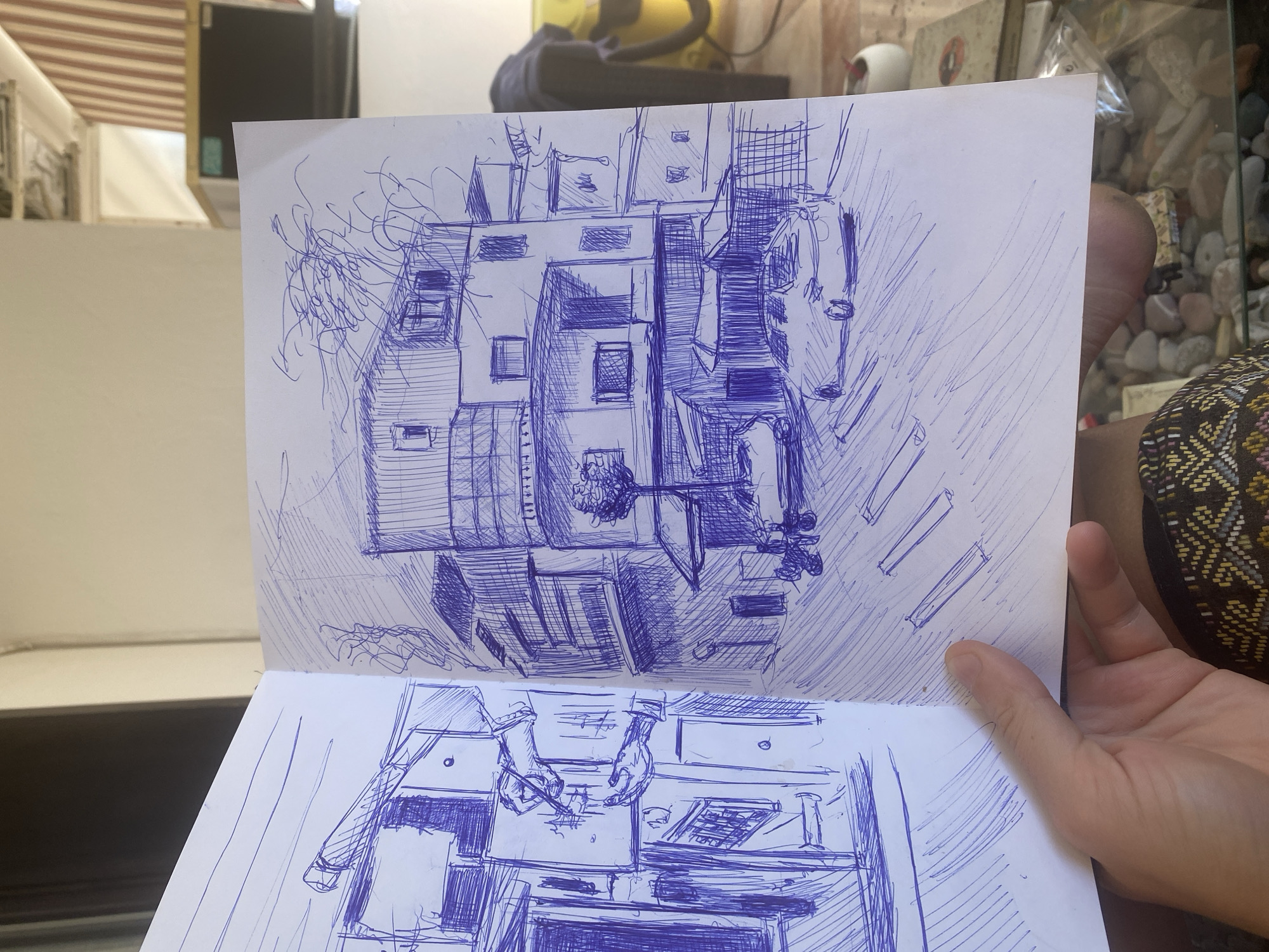 The image is a pen sketch on a sketchbook, showing an urban scene. The sketch is divided into two pages. The left page depicts a close-up view of buildings with windows and balconies, rendered with bold, intersecting lines. The right page shows a wider view of a street with multi-story buildings, a tree, two cars, and some people. The architecture suggests a European city, possibly Mediterranean, given the style of the buildings and the presence of a car and a tree on a street. The sketch is done in blue ink. In the foreground, a hand is holding the sketchbook. The background shows a blurred interior space with furniture and a window. The scene appears to be a street in Budva, Montenegro, with buildings lining the street and cars parked or driving. The sketch captures the essence of a bustling urban environment.
