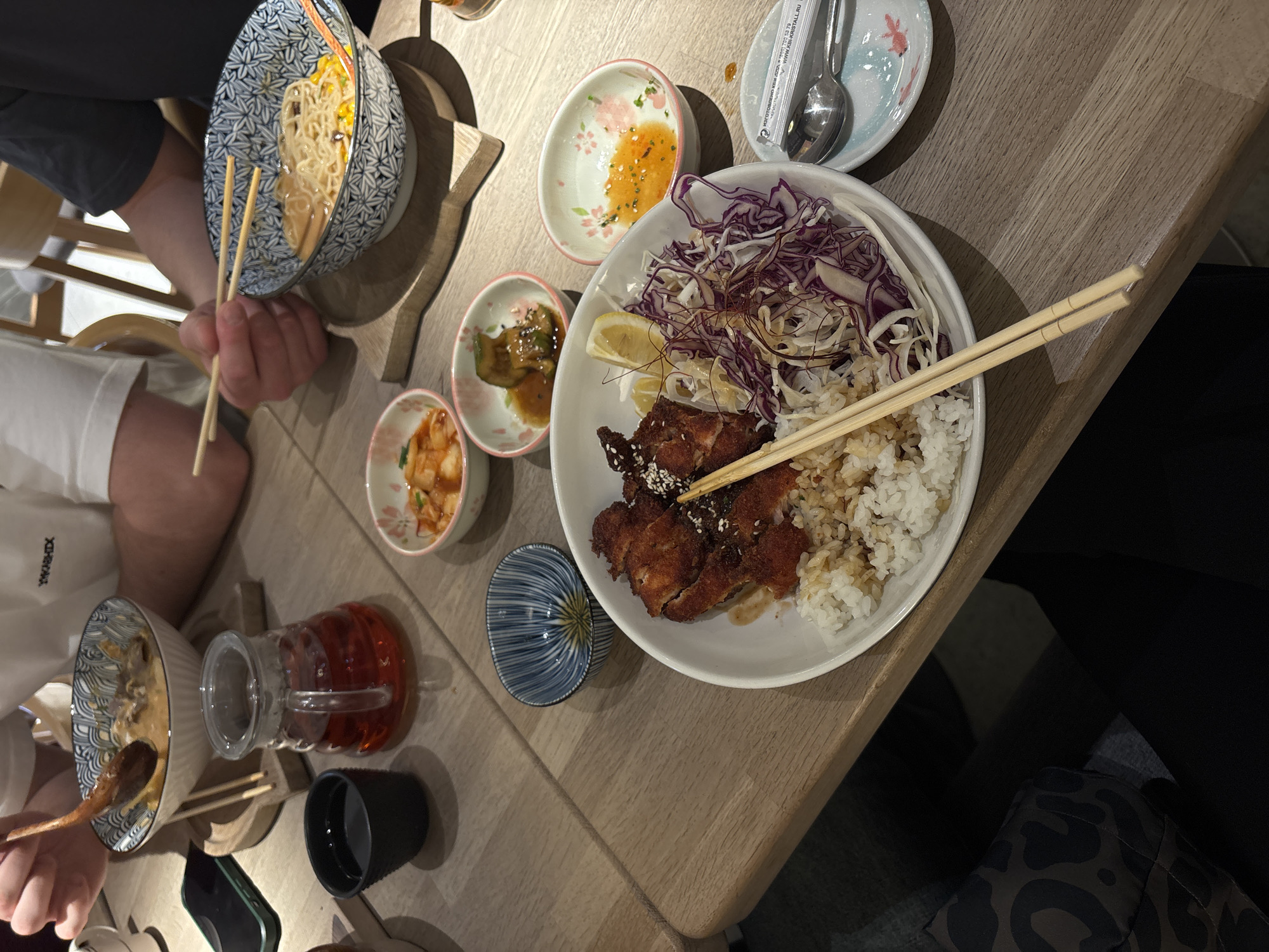 A high-angle shot shows a table setting with multiple bowls and dishes of Asian food, indicative of a dining experience. Two people are visible, each holding chopsticks and appearing to eat or prepare to eat the food. The table has a light-colored wood grain surface. On the table are bowls of noodles, rice with fried chicken and shredded purple cabbage, small side dishes of pickled vegetables, and sauces. A glass pitcher of a reddish-brown liquid and a small black cup are also present. The location is assumed to be in Moscow, Russia, based on contextual clues not explicitly visible in the image. The environment suggests a casual restaurant setting.