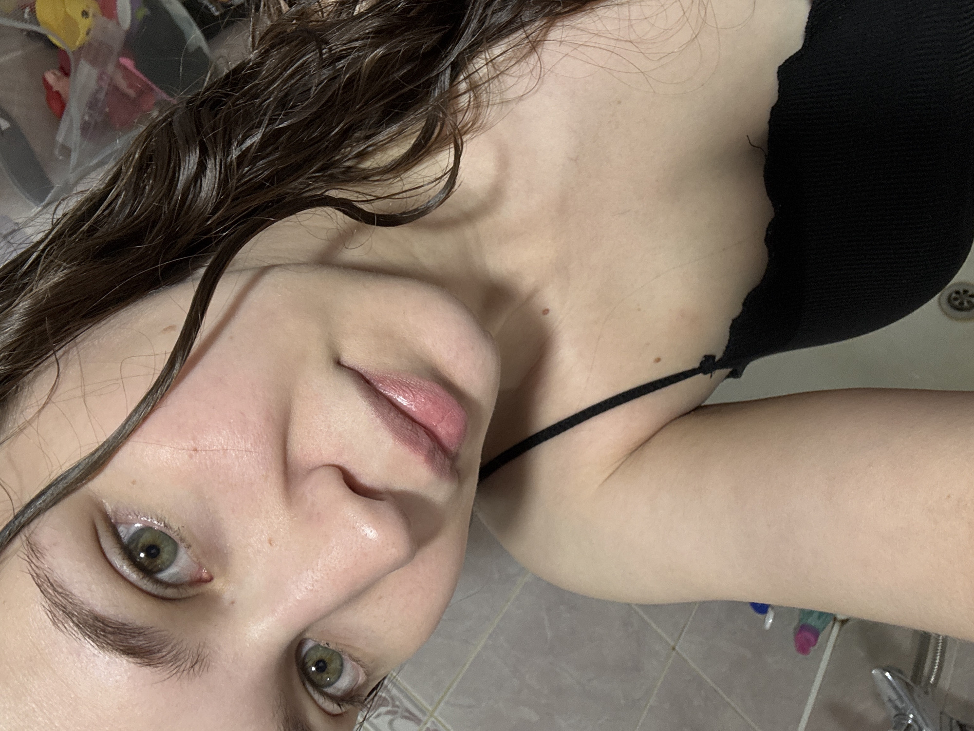 A close-up selfie of a young woman with wet, dark brown hair and light skin. She has prominent eyebrows, green eyes, and pale pink lips. She is wearing a black camisole. The background appears to be a bathroom, with tiled walls and shower fixtures visible. There are some indistinct objects in the background, including what appear to be cleaning supplies and possibly toys in a plastic bag. The lighting is soft, and the image is taken from a slightly elevated angle. There is no discernible location in Chaikovsky, Russia, evident in the image.