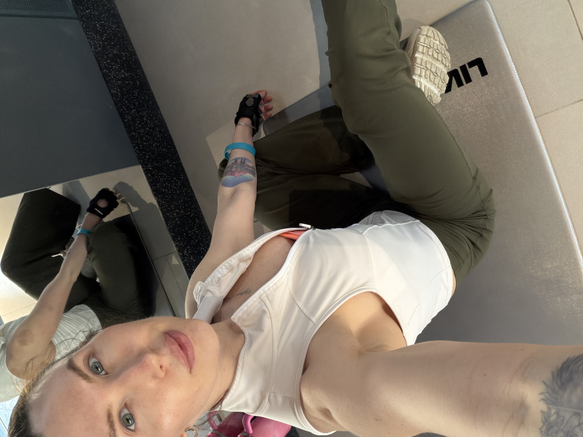 A woman takes a selfie indoors. She is seated on a gray mat with black lettering "LIV". She wears green pants, a white top, and a black glove on her left hand. She has tattoos on her arms. Her right foot, in a white sneaker, is on the mat. A mirror is partially visible on the left side of the frame. The location is presumed to be indoors, possibly in Perm, Russia. No specific details link the image to Perm or a specific location within the city.