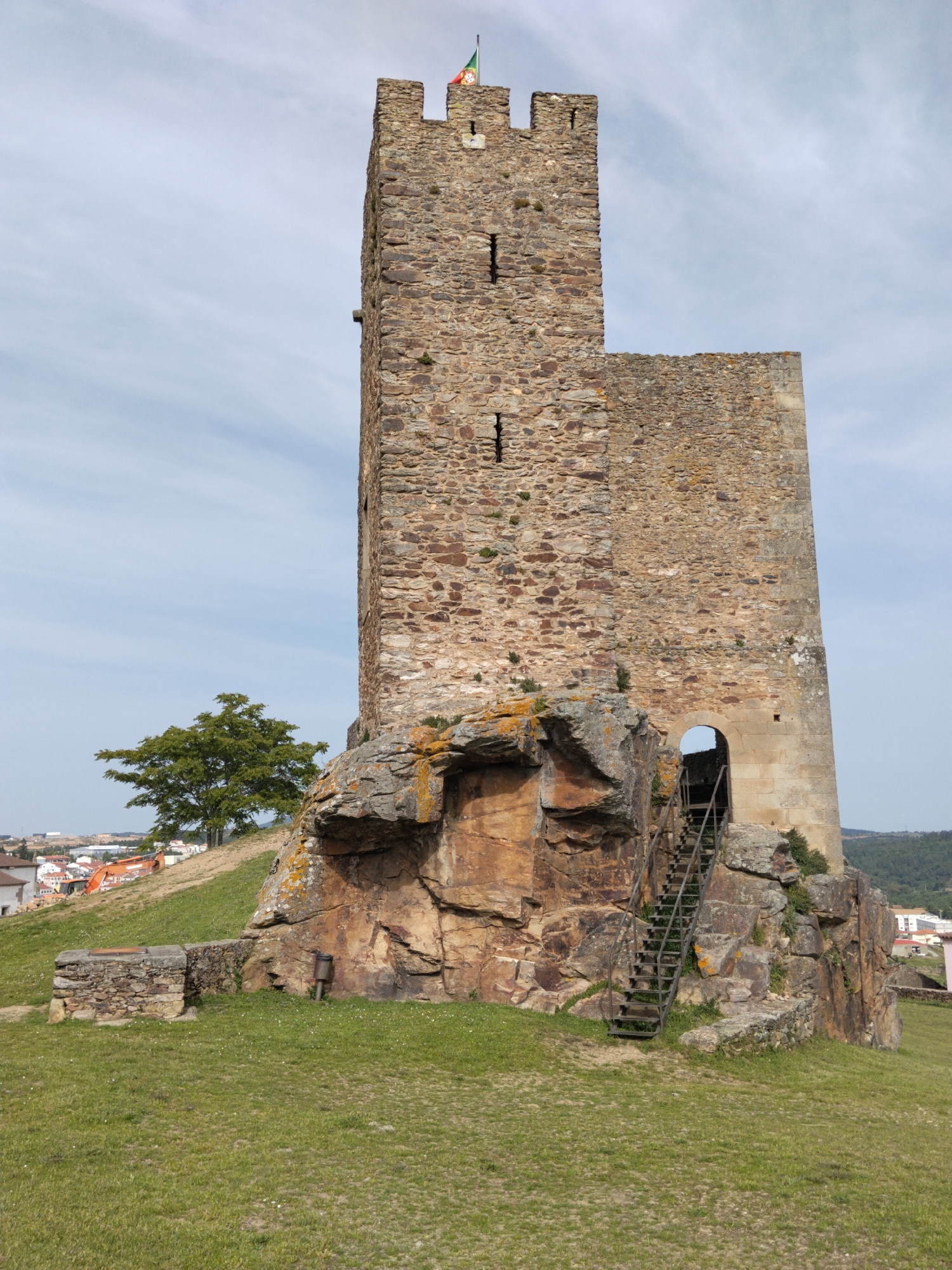 The image shows the Penas Roias Castle tower, located in Mogadouro, Portugal. The tower is made of stone and sits atop a rocky outcrop. A set of metal stairs leads up to an arched entrance in the side of the tower. A green flag flies from the top of the tower. The surrounding environment consists of a grassy field and a tree on the left, with parts of Mogadouro visible in the background.