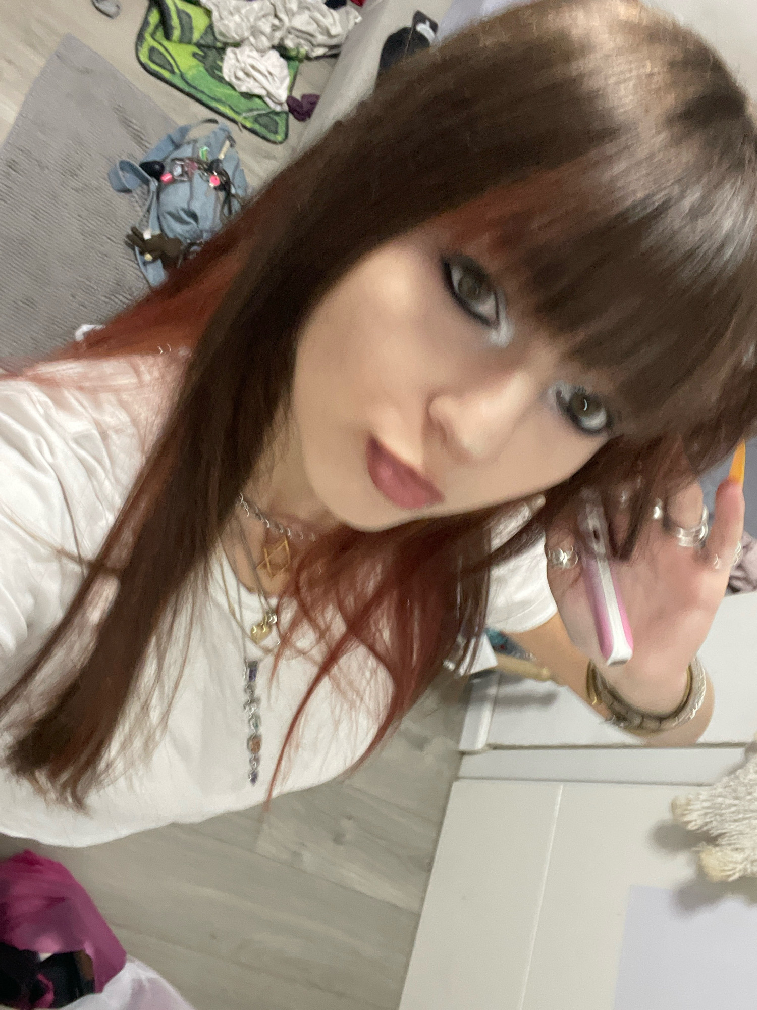 The image shows a close-up, high-angle shot of a young person with long, brown hair with red undertones and bangs, wearing a white t-shirt and multiple necklaces. The person is wearing heavy eye makeup with prominent eyeliner and eyeshadow. They are holding a pink and white object, possibly a vape or a small electronic device, in their right hand, which also features several rings and brightly colored nail polish. The background is blurry but appears to be an indoor setting with a gray rug, some clothing items on the floor, and white furniture. The scene does not contain any identifiable landmarks or elements that would indicate the location is Raanana, Israel.