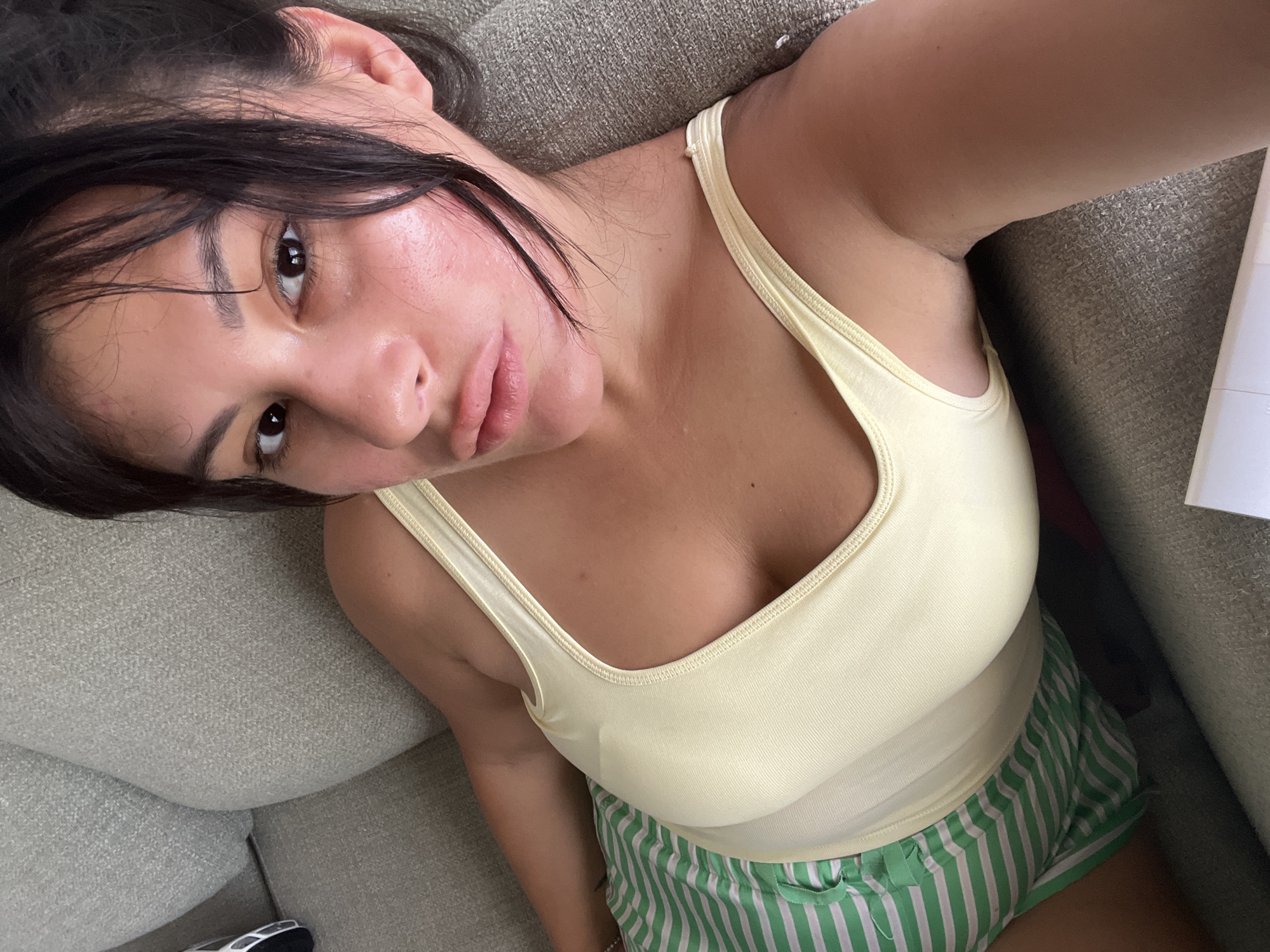 The image shows a young woman lying on a light-colored couch. She is looking up towards the camera, with her head tilted to the side. Her dark hair is loose and falls around her face. She is wearing a pale yellow tank top and green and white striped shorts. The room appears to be indoors, with the couch being the dominant element of the setting. The lighting suggests it is daytime. The woman's expression is relaxed, and she appears to be in a casual, comfortable setting. There is no visible text in the image. The location is Kiryat Bialik, Israel.