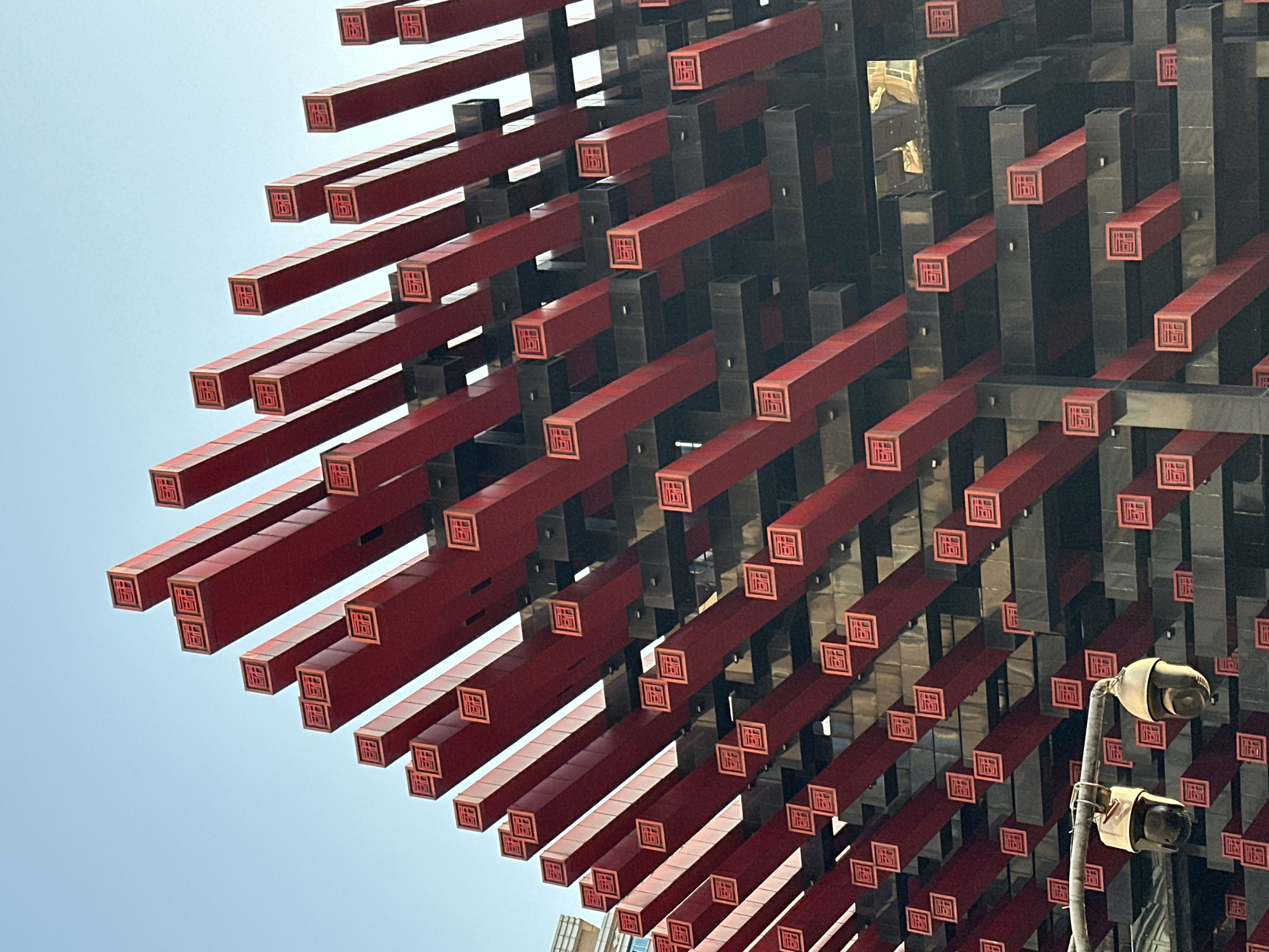 The image depicts a portion of the Raffles City Chongqing complex in Yuzhong District, China. The structure features a grid-like design composed of red and black rectangular beams, each adorned with a red square containing a Chinese character. The beams extend in varying lengths, creating a multi-dimensional pattern against a light blue sky. A surveillance camera mounted on a pole is visible in the lower right portion of the image.