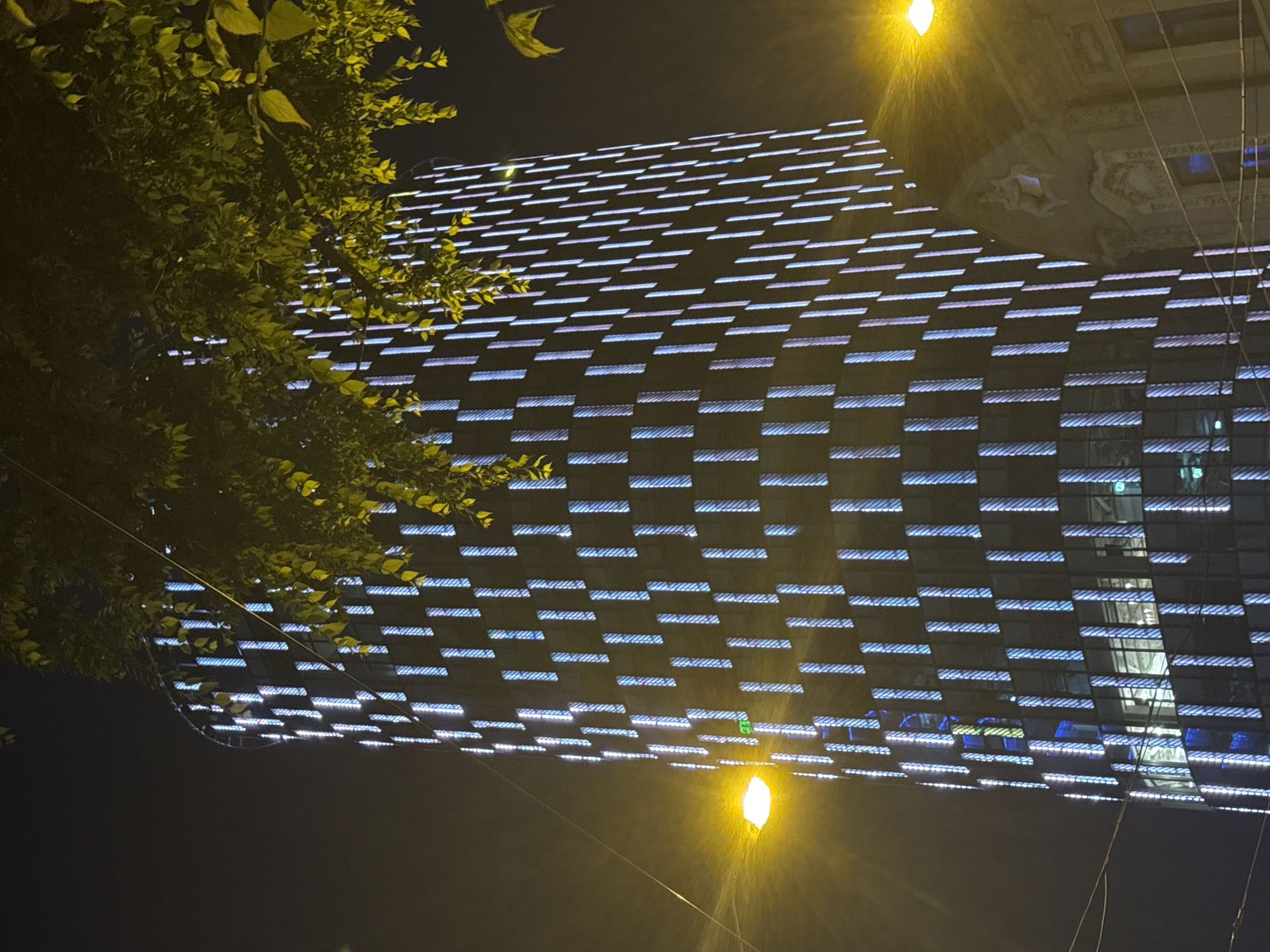 The image depicts a nighttime scene of the National Bank of Slovakia building in Bratislava, Slovakia. The skyscraper's facade is illuminated with vertical LED lights in alternating shades of blue and purple, creating a striking visual effect against the dark sky. Trees with yellow-green leaves are visible in the foreground, partially obscuring the top of the building and adding a natural element to the urban landscape. Streetlights cast a warm, yellow glow, contributing to the overall ambiance. The building's modern architecture is characterized by its cylindrical shape and the repeating pattern of illuminated vertical elements. A portion of an older building with ornate architectural details is visible to the right of the skyscraper. The scene is characterized by artificial lighting and the stark contrast between the illuminated skyscraper and the dark, night sky.