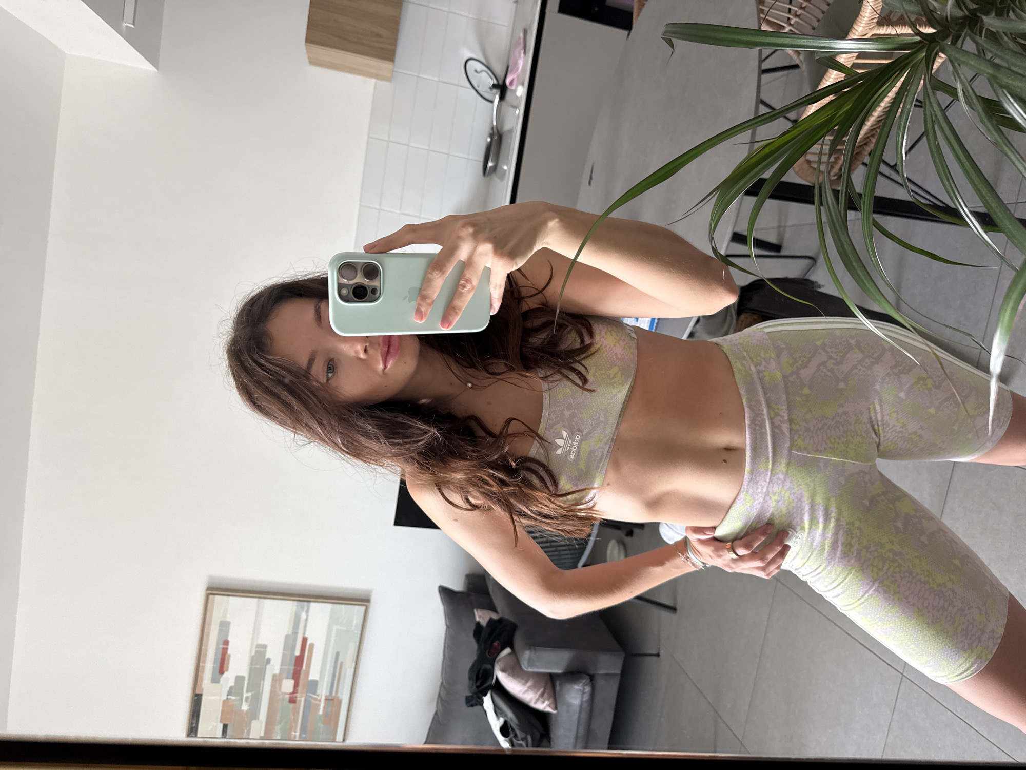 A young woman with brown hair is taking a selfie in a mirror. She is wearing a matching Adidas sports bra and biker shorts with a light green and grey snakeskin pattern. She is holding a light green iPhone. The reflection shows a modern apartment interior with a grey couch, a piece of art on the wall, and a kitchen area with white tile. A potted plant is partially visible in the foreground. The location is likely Givatayim, Israel, based on the architectural style and common interior designs of apartments in the area.