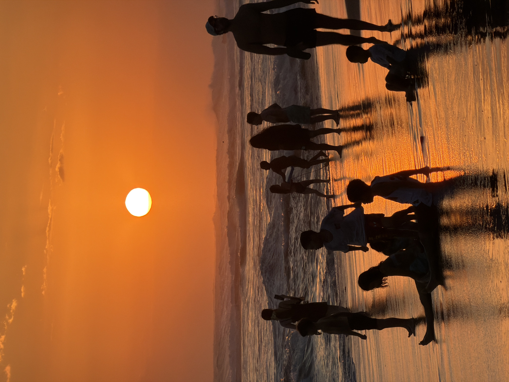 This is a vertical, eye-level shot of a beach in North Kuta, Indonesia, during sunset. The sun is a bright, white orb in the upper center of the frame, casting a warm orange glow across the sky and the ocean. The water has moderate waves, reflecting the sunset. Silhouetted figures of people, appearing to be mostly children and a few adults, are scattered across the wet sand and in the shallow water. Some are standing, some are sitting, and some appear to be playing. The wet sand reflects the orange light, creating long shadows. The overall mood is peaceful and idyllic.