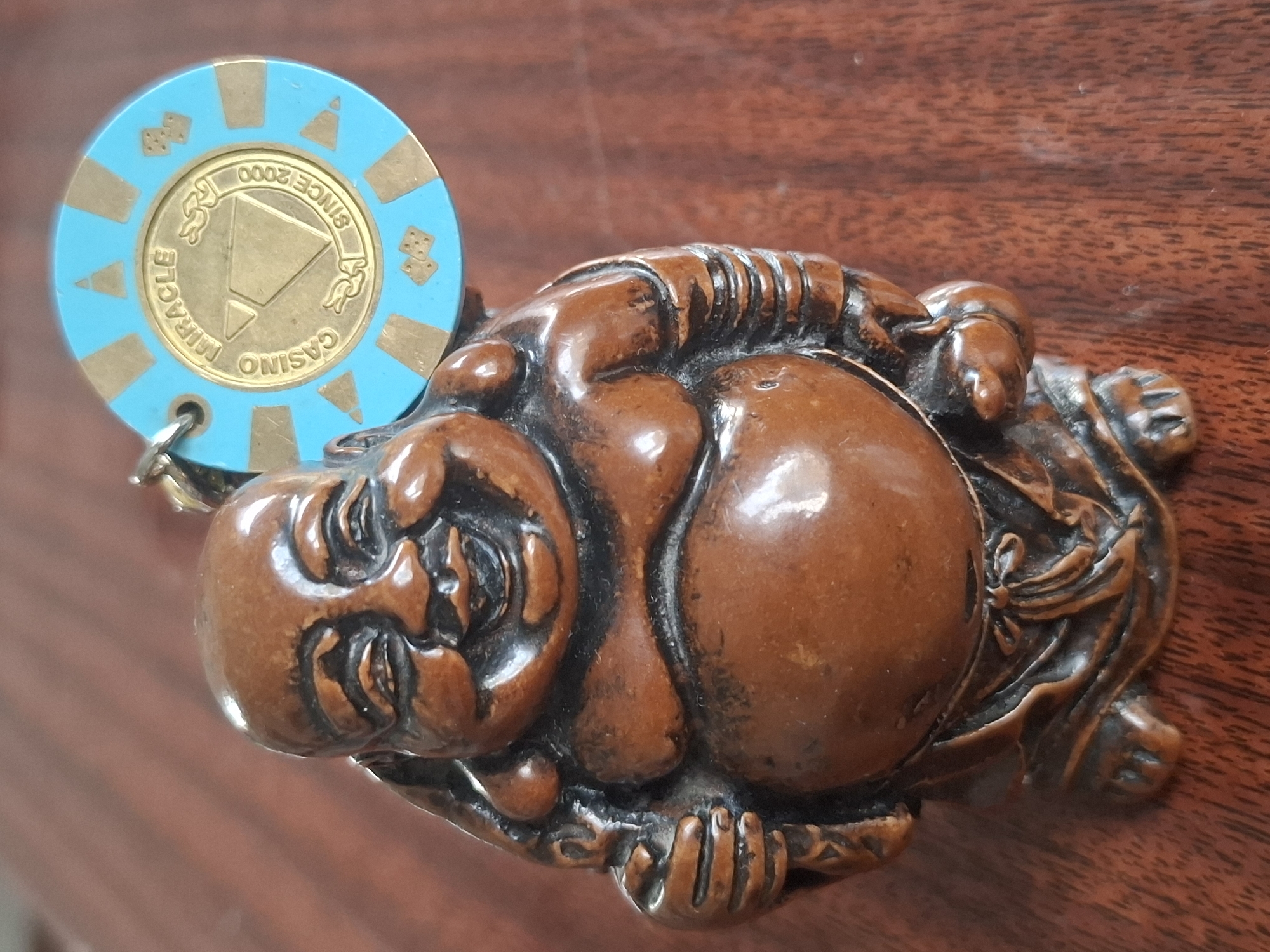 The image shows a close-up of a small, brown, carved figurine of a laughing Buddha. The figurine is intricately detailed, with folds of clothing and a jovial expression. Attached to the figurine by a small metal loop is a round, blue and gold casino chip. The chip has a gold inset that reads "CASINO MIRACLE" in a circular pattern, with a diamond shape in the center. Around the inset, it says "SINCE 2000". The chip also features small gold triangle shapes and dice symbols on its blue surface. The background is a blurry, reddish-brown wooden surface, likely a table. The scene is indoors, with warm lighting. There is no information about the location in Almaty, Kazakhstan.
