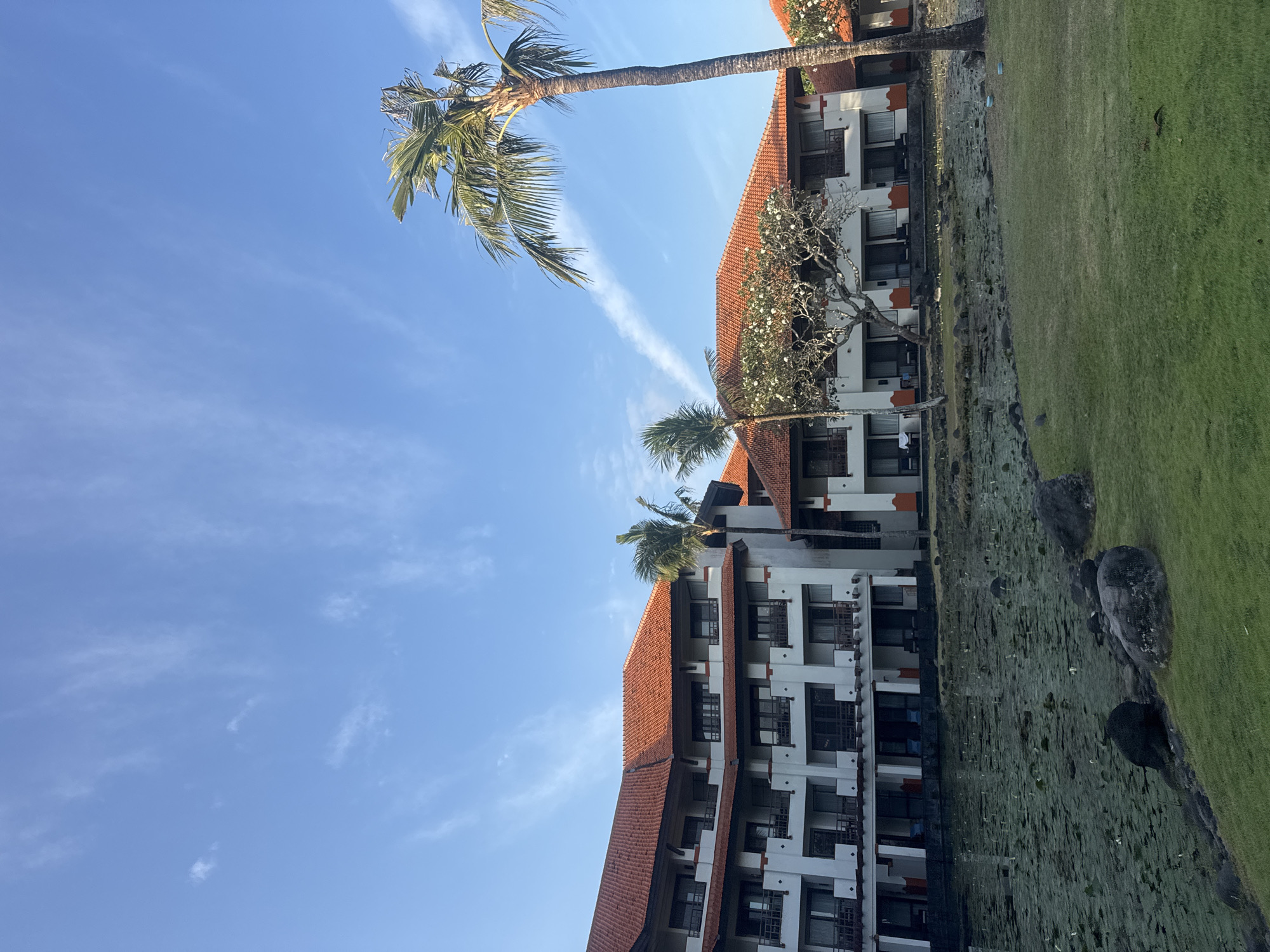 This image captures a serene view of a resort in Kuta Selatan, Indonesia, on a clear day. A large, multi-story building with red tiled roofs dominates the scene, its white facade adorned with balconies and dark-framed windows. In the foreground, a calm pond is covered with lush green lily pads, with a few dark rocks scattered along its edge. A meticulously manicured lawn stretches out from the pond's edge towards the resort. Several palm trees, including a tall, slender one on the right, punctuate the landscape, their fronds swaying gently in the breeze. The sky above is a vibrant blue, streaked with thin, wispy clouds, suggesting a peaceful atmosphere. The overall impression is one of tranquility and tropical beauty.