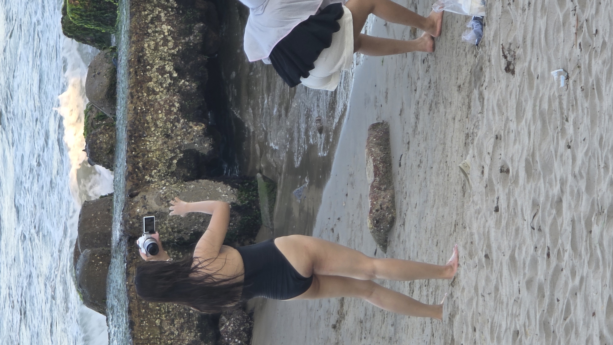 The image shows a beach scene in Nha Trang Ward, Vietnam. A woman in a black bathing suit stands on the sand near a rocky breakwater, holding a camera to take pictures of the waves crashing against the rocks. Another person in a white top and black skirt is also standing on the sand further away. The beach is sandy with some scattered debris.