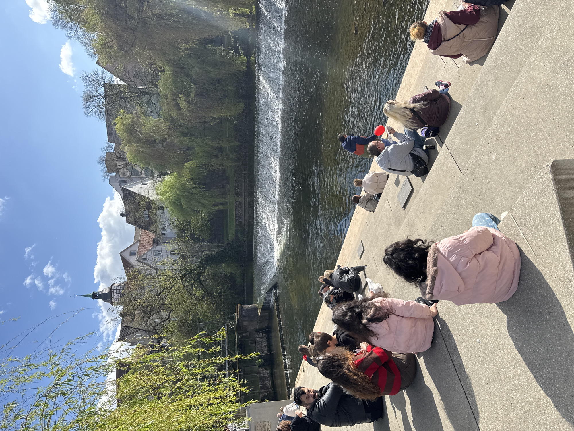 The image depicts a group of people sitting on concrete steps adjacent to a river with a small waterfall in Backnang, Germany. The setting suggests a warm, sunny day.
Several people are seated on the steps facing the water. Many people are wearing jackets. A person is holding a red balloon.
Across the river is a combination of greenery (trees) and old buildings including buildings with a steeple. The sky is blue with some scattered clouds. The overall scene appears to be a relaxed, public space near a scenic waterway.
