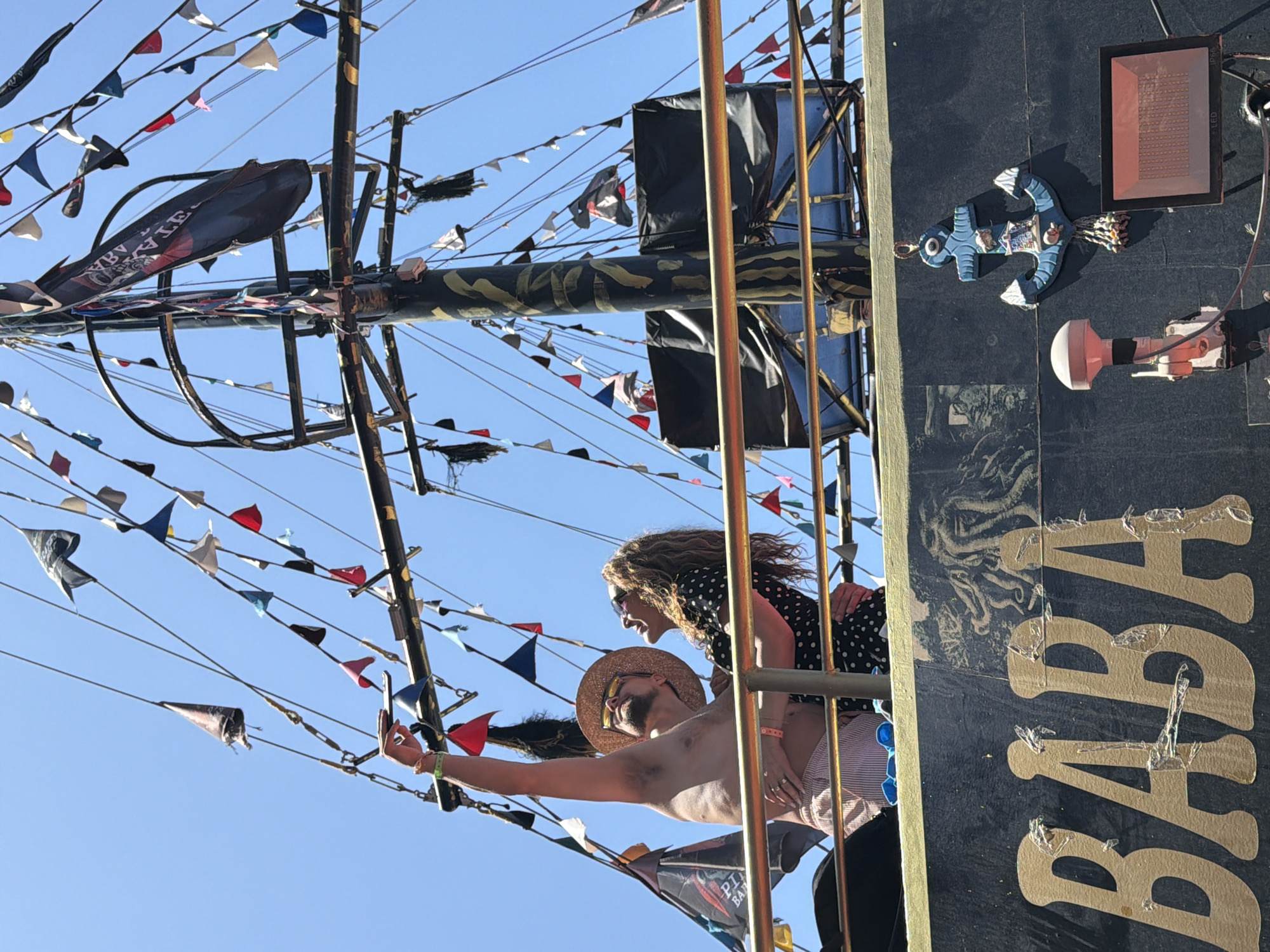 This vibrant image captures two individuals, a man and a woman, enjoying themselves on what appears to be a pirate-themed boat or ship, under a clear, bright blue sky. The scene is illuminated by strong daylight, indicating good weather.
The man, shirtless and wearing a straw hat and sunglasses, is in the foreground, stretching his arm out to hold a smartphone, likely taking a selfie or video. He has a joyful expression. Behind him, the woman, with curly hair and sunglasses, wearing a polka-dot top, smiles broadly as she holds onto a metal railing. Both convey happiness and leisure.
They are positioned within a section of the ship's rigging or a crow's nest, which is elaborately decorated. Numerous small, triangular flags (bunting) in various colors are strung along the lines and masts, adding a festive and celebratory atmosphere. Larger black fabric elements, possibly decorative sails or coverings, are also visible. On a black banner hanging from the rigging, part of the word "PIRATE" can be seen.
To the right, the dark side of the ship features the prominent gold letters "BABA" spelled vertically. Above this text, a decorative light blue anchor is affixed, alongside a rectangular black LED light fixture and another smaller white light.
The overall setting suggests a tourist attraction or a themed boat tour, consistent with the Alanya, Turkey location, where such pirate ship excursions are popular.