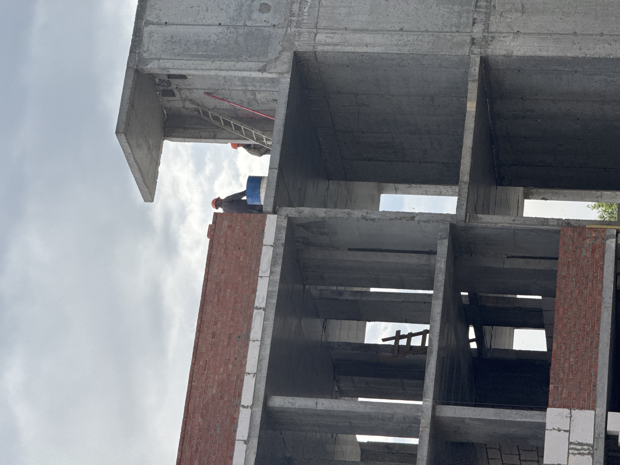 A construction site in Izhevsk, Russia is depicted from a low-angle view. The concrete and brick framework of a multi-story building is under construction. Two construction workers wearing hard hats are visible on the upper floors. One worker is carrying a blue barrel. Ladders are positioned within the structure for access to the different levels. The sky is overcast.