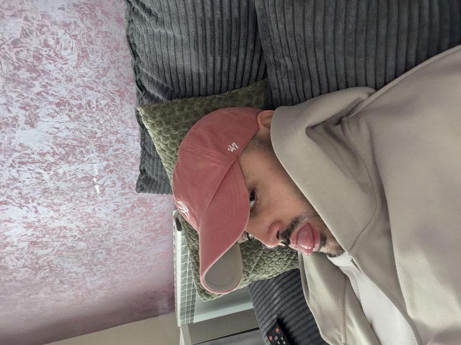 A male individual is depicted reclining in an indoor setting, identified as Sofia, Bulgaria. He wears a light beige hooded jacket over a white ribbed shirt and a faded pink baseball cap with a "47" logo embroidered on the right side. The male has a dark mustache and beard, and his tongue is extended from his mouth. He is positioned against a gray ribbed couch, with a textured green cushion and another gray ribbed cushion visible behind him. A black television remote control rests on the couch to the left of the individual. The background wall features a textured, metallic pink wallpaper pattern. A white radiator is partially visible in the lower left portion of the image.