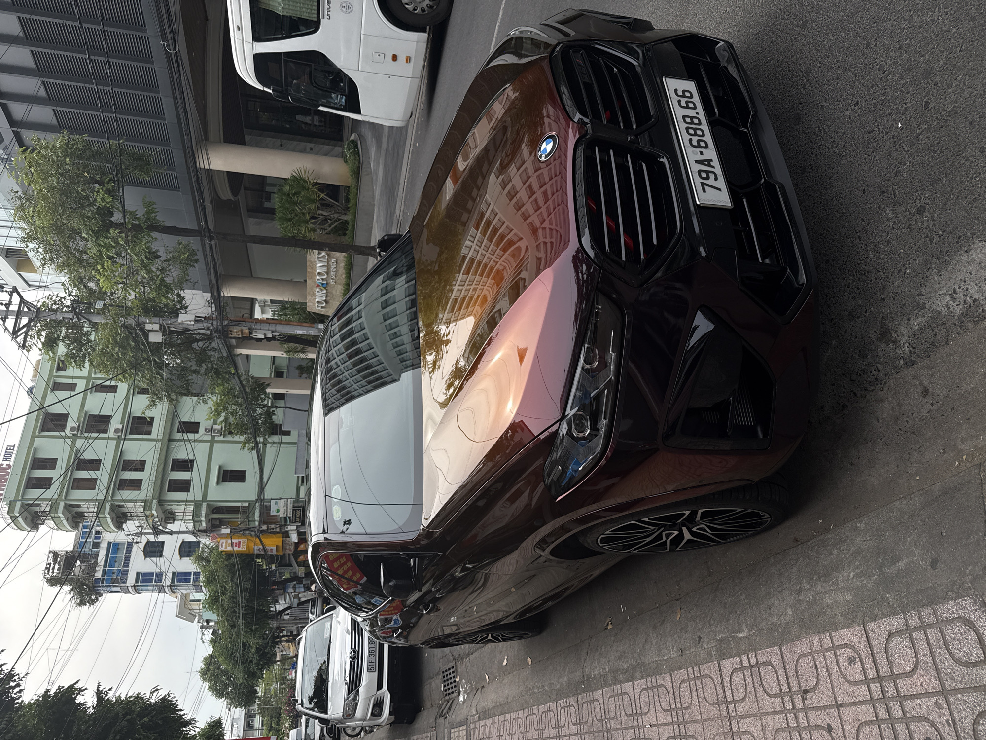 The image shows a luxury BMW X6 automobile parked on a sidewalk in Nha Trang Ward, Vietnam. The car has a dark red-brown color with a prominent BMW logo on the front grille. Its license plate reads "79A-688.66". The car reflects the surrounding buildings due to its polished surface. The sidewalk is paved with patterned tiles. In the background, there's a white van and a multi-story building. Street foliage and electrical wires are also visible in the background, common features of urban scenes in Vietnam.