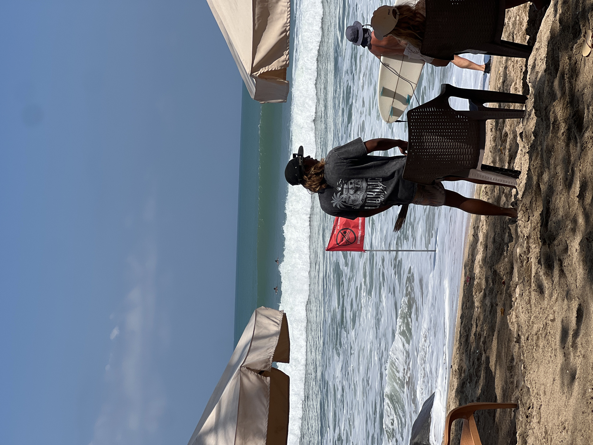 This image depicts a beach scene in Kuta, Indonesia, likely during daytime. In the foreground, a sandy beach is visible with shadows cast by unseen umbrellas. Two brown plastic chairs are positioned on the sand. To the right of these chairs, a person with long brown hair, wearing a black t-shirt and a black baseball cap, stands facing away from the camera, looking towards the ocean. They appear to be holding a broom. A red flag with a white symbol is planted in the sand near the water's edge, indicating "SWIMMING PROHIBIT."
Further back, a white surfboard is leaning against a person wearing a dark hat and an orange shirt, also facing away from the camera. More people are visible in the ocean, appearing to be surfers. The ocean has moderate waves with white foam breaking near the shore. In the background, the ocean stretches out to the horizon under a clear blue sky with a few wispy clouds. Two large, light-colored beach umbrellas are partially visible on either side of the frame, providing shade to areas of the beach. The overall atmosphere suggests a typical beach day in a popular tourist destination.