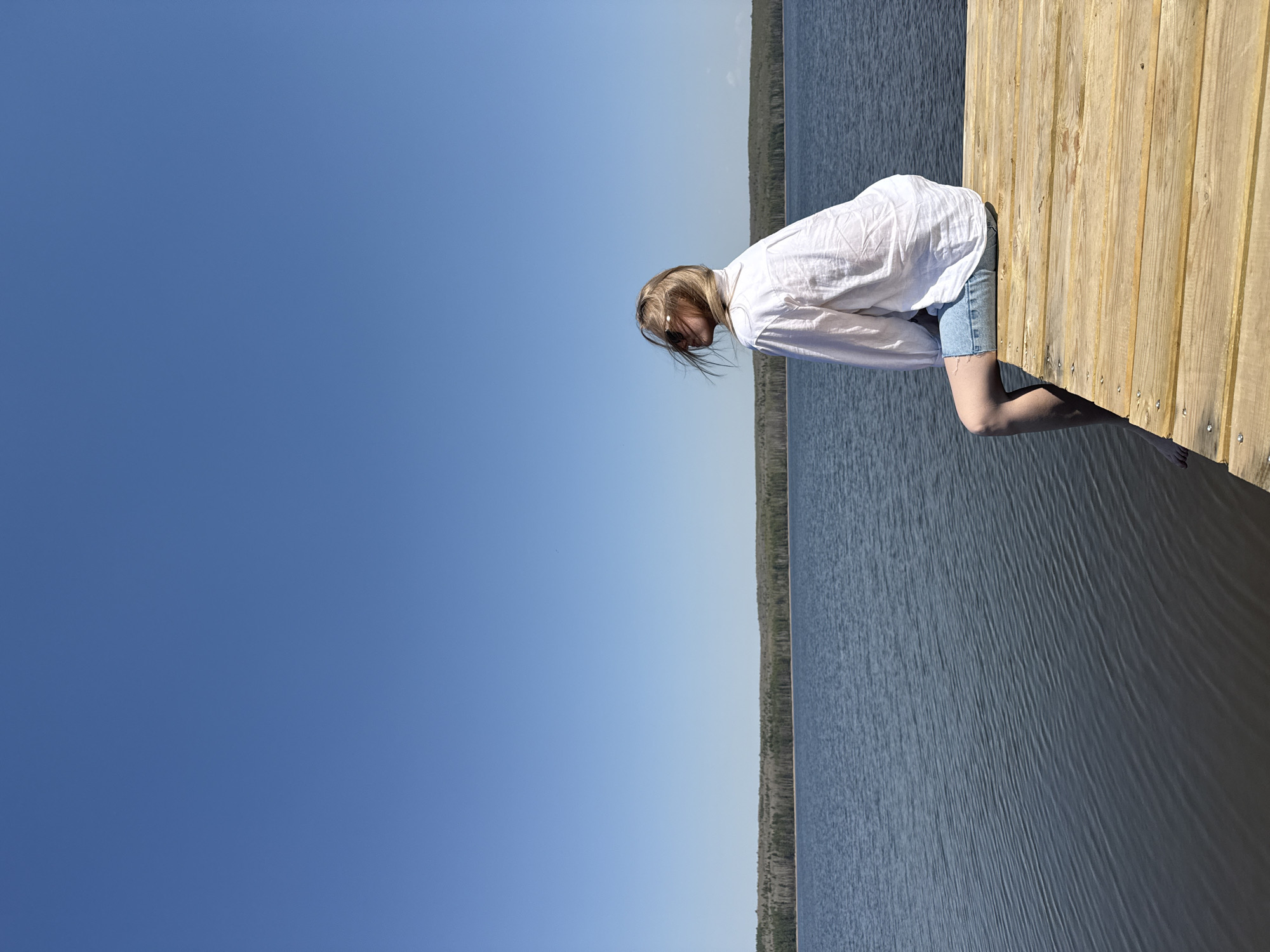 A young woman stands on a wooden dock or pier extending into a body of water, possibly a lake or river. She leans forward with her hands resting on the edge of the dock. She has fair hair and is wearing a white shirt and denim shorts. The water stretches to the horizon where a line of trees is visible. The sky is clear blue. The image appears to be taken in Chaikovsky, Russia.
