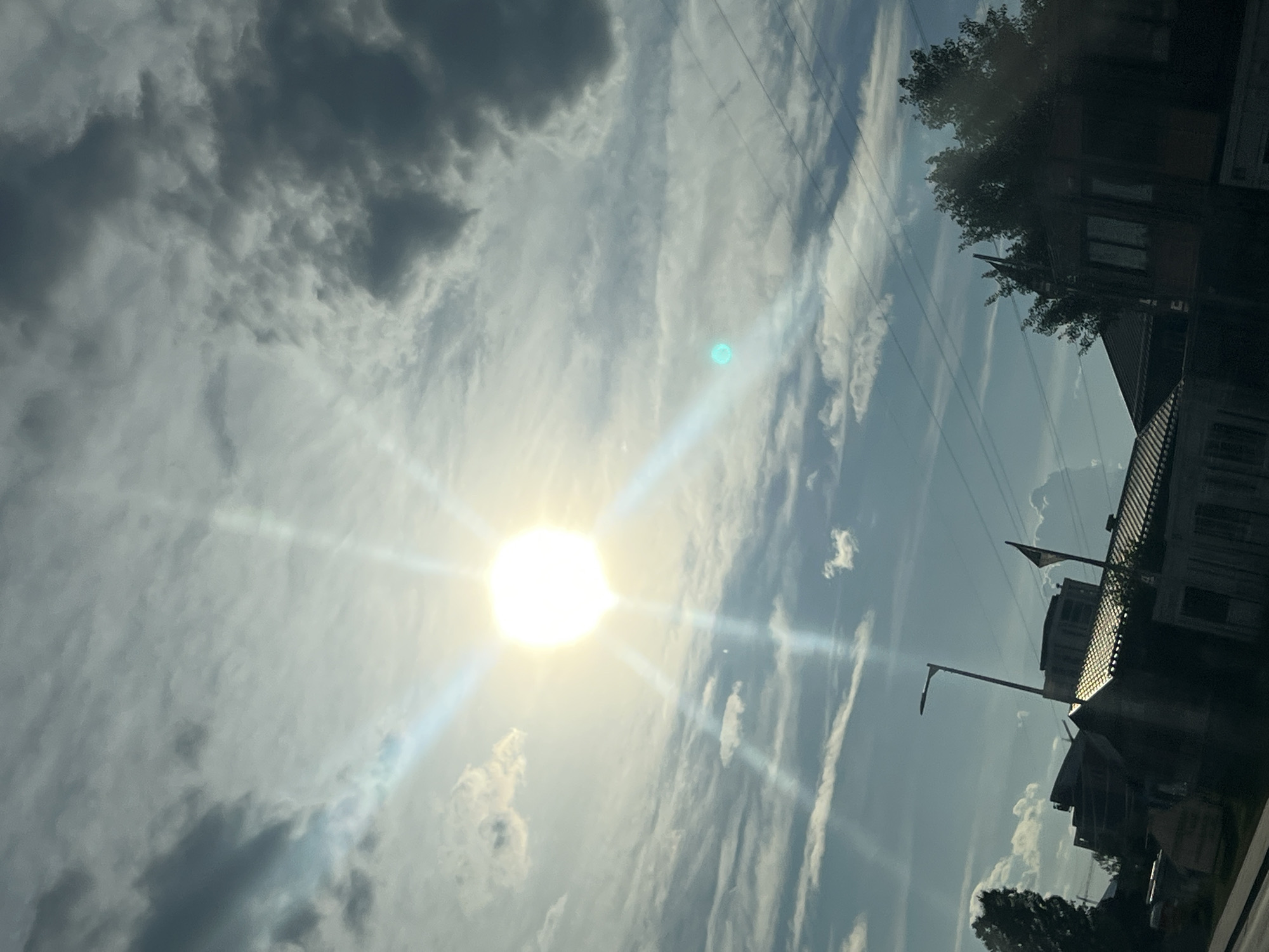 The image shows a bright sun with strong rays breaking through a cloudy sky. Some of the clouds are dark and ominous, while others are wispy and light. In the foreground, there are buildings and trees silhouetted against the sky. There are also power lines visible. The scene appears to be from a city, but the specific location is unknown.