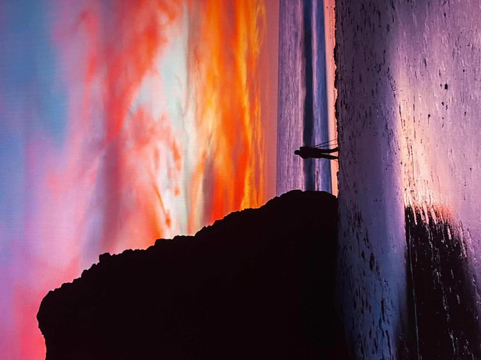 The image depicts a photographer silhouetted against a vibrant sunset over a beach. A large, dark rock formation is visible on the left side of the frame. The sky is a riot of color, with shades of pink, orange, and blue from the setting sun. The ocean is calm, with gentle waves lapping at the shore. The sand is wet and reflective, mirroring the colors of the sky. The photographer is standing behind a tripod, apparently capturing the scene. This image does not depict any location in the city of Krasnokamsk, Russia.