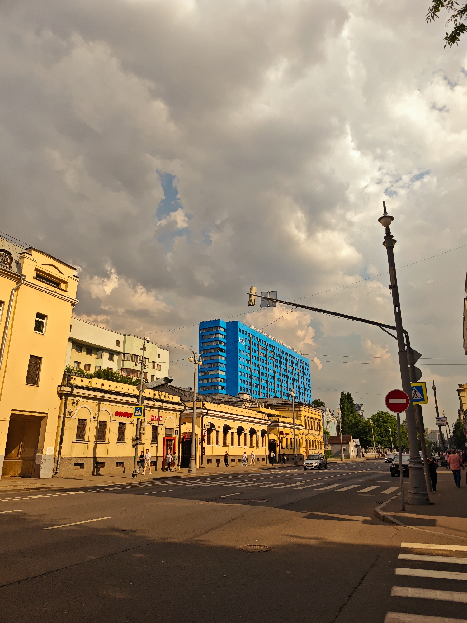 The image depicts a street scene in Moscow, Russia, under a dramatic, cloudy sky. In the foreground, a paved road with white crosswalk markings dominates the lower half of the frame. To the left, a yellow building with arched windows and a wide entryway is visible. Adjacent to it, a longer, low-rise yellow building with a dark roof features a series of windows and doors, with several people walking on the sidewalk in front of it. Further down the street, a striking, tall blue building with numerous windows stands out against the cloudy sky. A lamppost with traffic signals and signs, including a red circular "no entry" sign and a pedestrian crossing symbol, is positioned on the right side of the street. Several cars are visible on the road, and more pedestrians are present on the sidewalks on the right. The overall impression is a typical urban environment with a mix of architectural styles, caught in soft, late afternoon light.