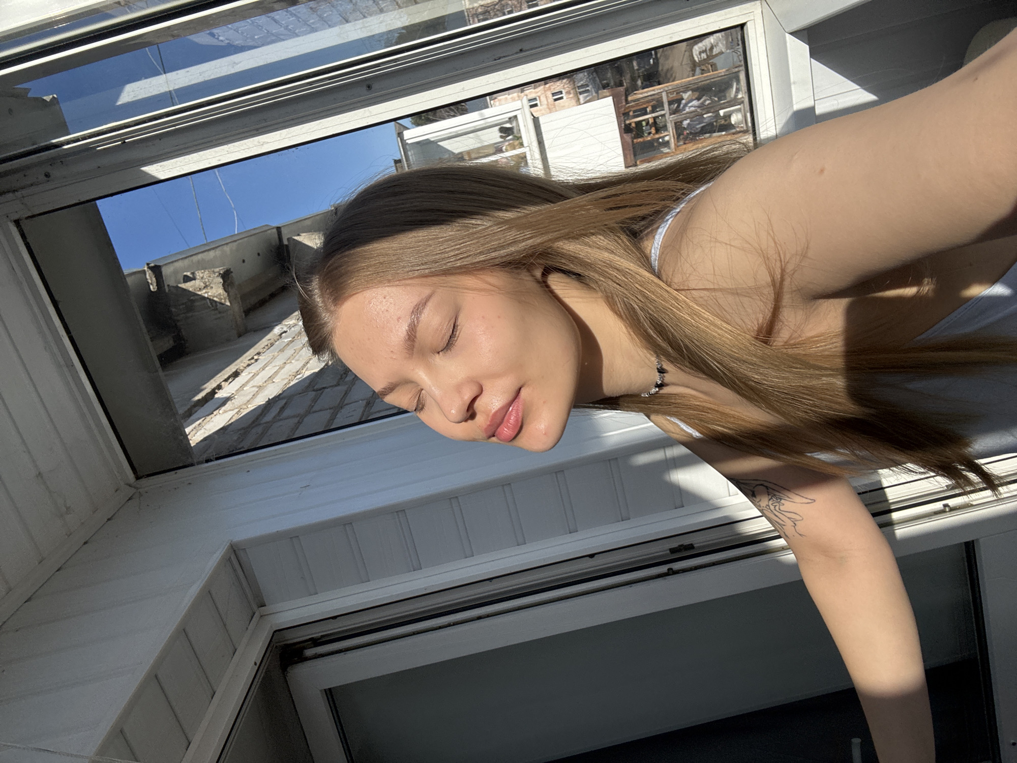 A young woman with light brown hair and closed eyes is the central subject, appearing to be resting or sunbathing. Her head is tilted upwards, and her lips are slightly parted in a relaxed expression. She wears a light-colored top and a necklace with dark beads. A linear tattoo of a cartoon character, resembling Bugs Bunny, is visible on her left upper arm.
The woman is positioned near large windows, likely on a balcony or in a room with a bright outlook. The immediate surroundings include white paneled walls and window frames. Beyond the windows, a clear blue sky dominates, indicating bright and sunny weather. Distant urban architecture, including building facades and rooftops, can be seen, partially reflected in the windowpanes. The strong, natural sunlight casts clear shadows, signifying it is daytime.
The scene conveys a sense of peace and relaxation, with the woman seemingly enjoying the warmth and light. The image provides visual cues of an urban setting under fair skies, with the location context being Chaikovsky, Russia. No discernible text is visible within the image.